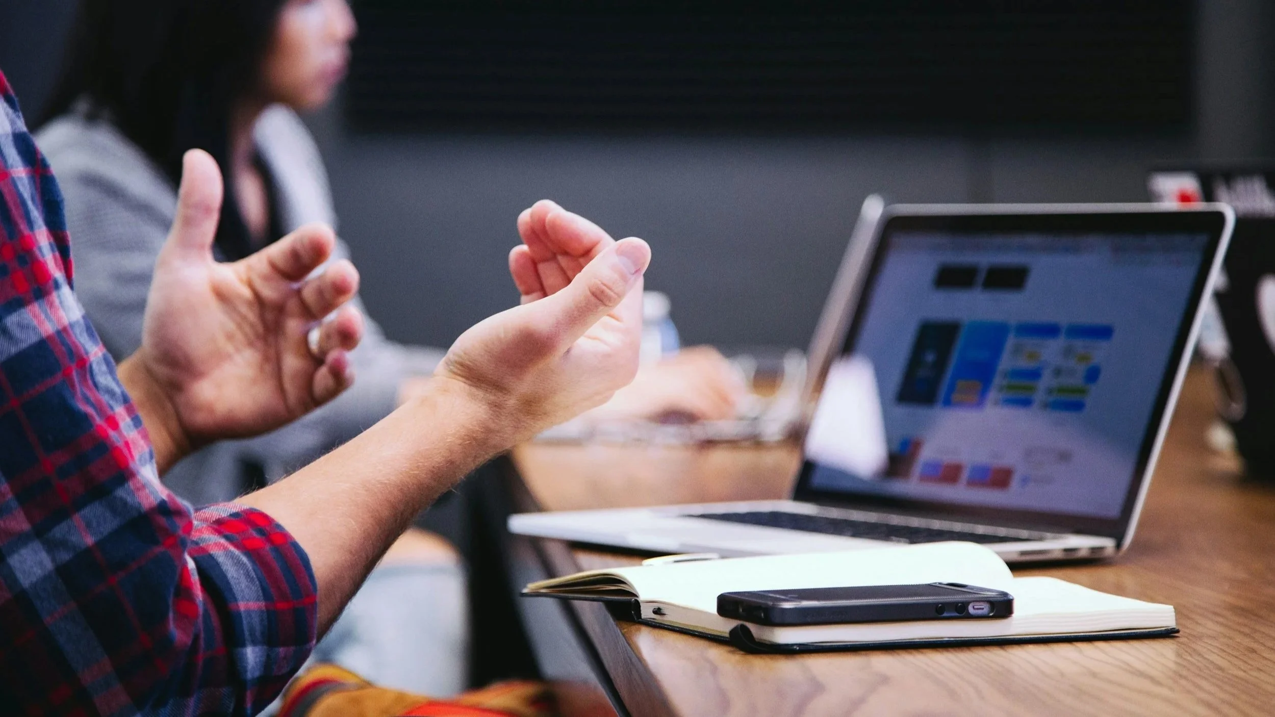A person in a plaid shirt gesturing with their hands while talking during a meeting, with a woman in the background working on a laptop at a wooden table.