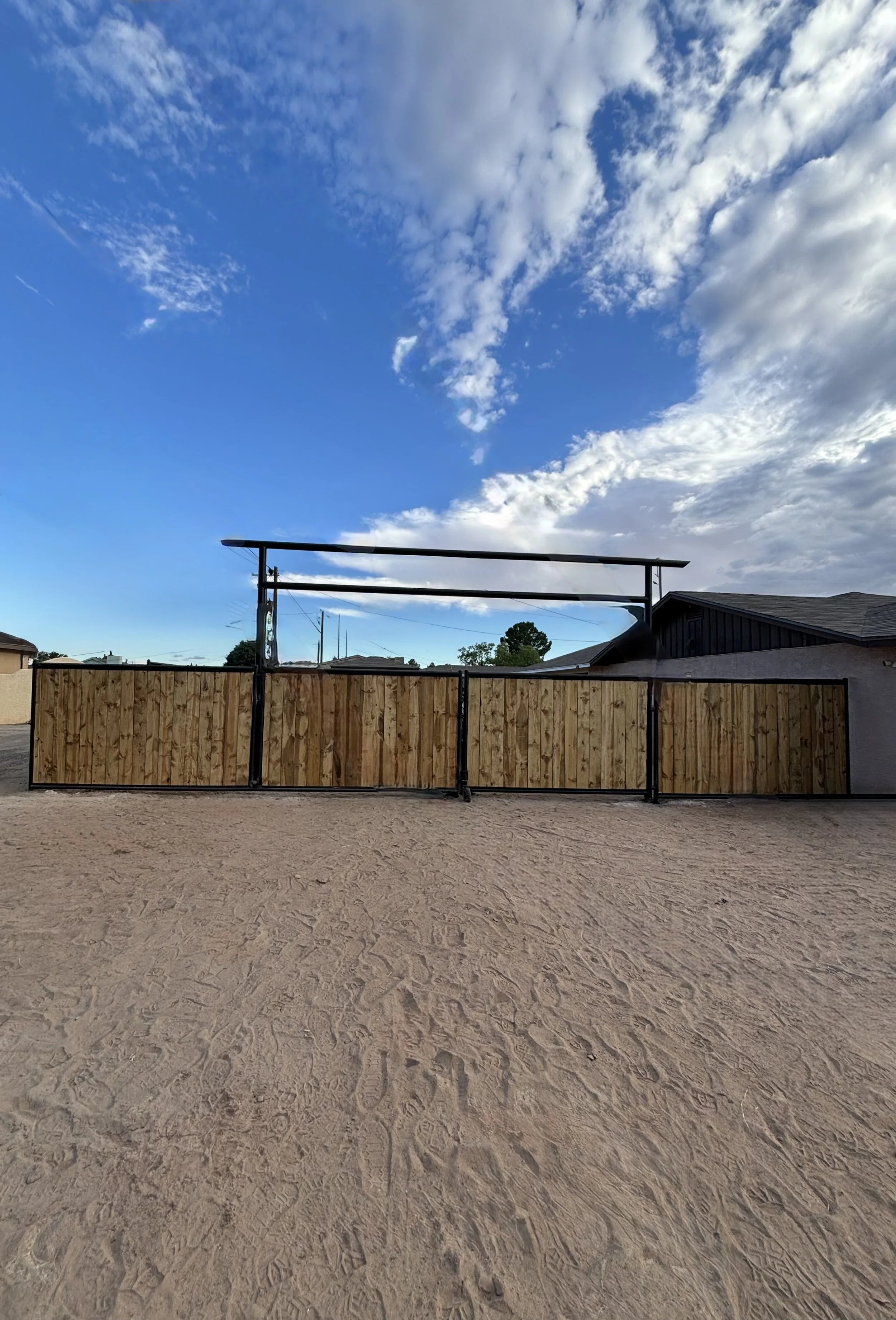 A sandy ground with some footprints in front of a wooden fence and metal framework, under a sky with scattered clouds and a mix of blue and white sky.