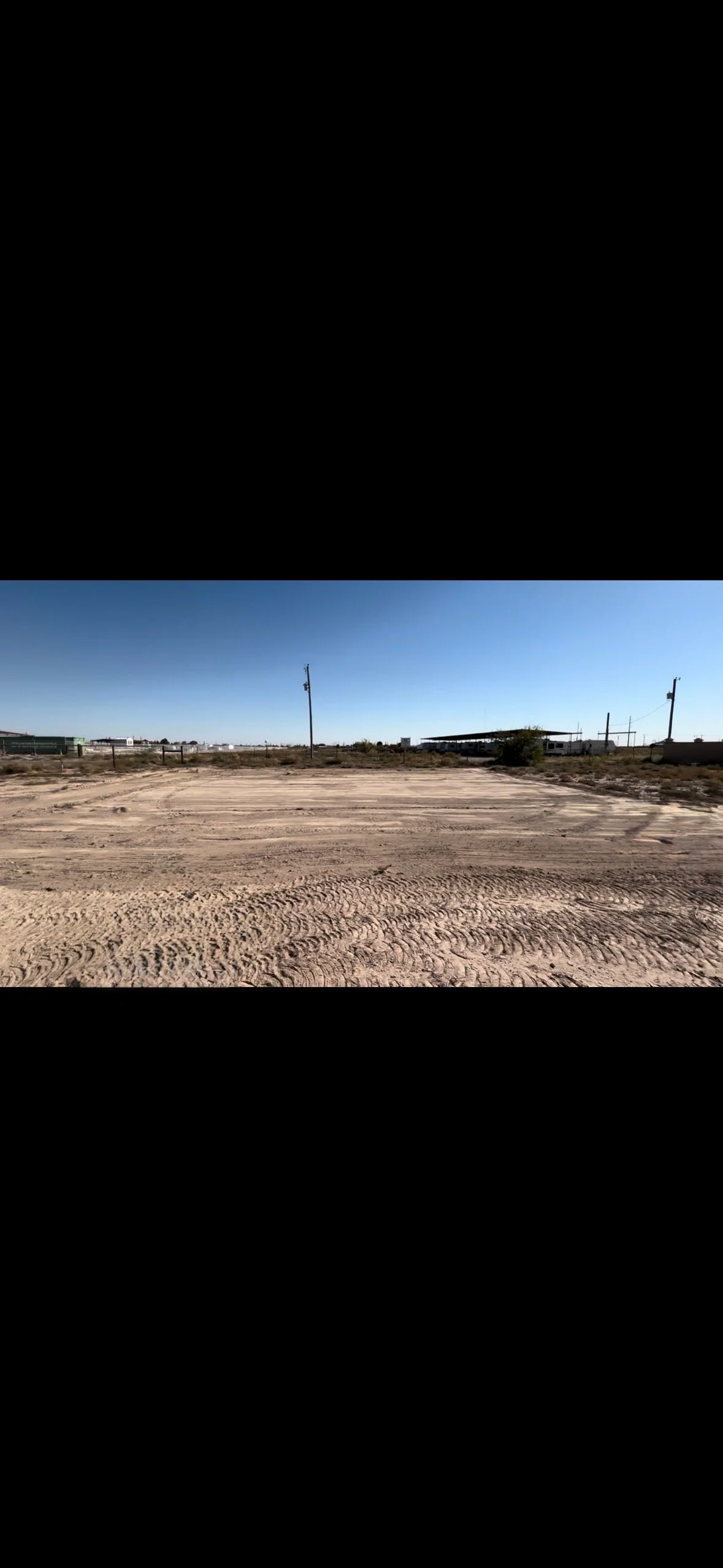 An empty dirt lot under a clear blue sky, with a few power poles and distant buildings.