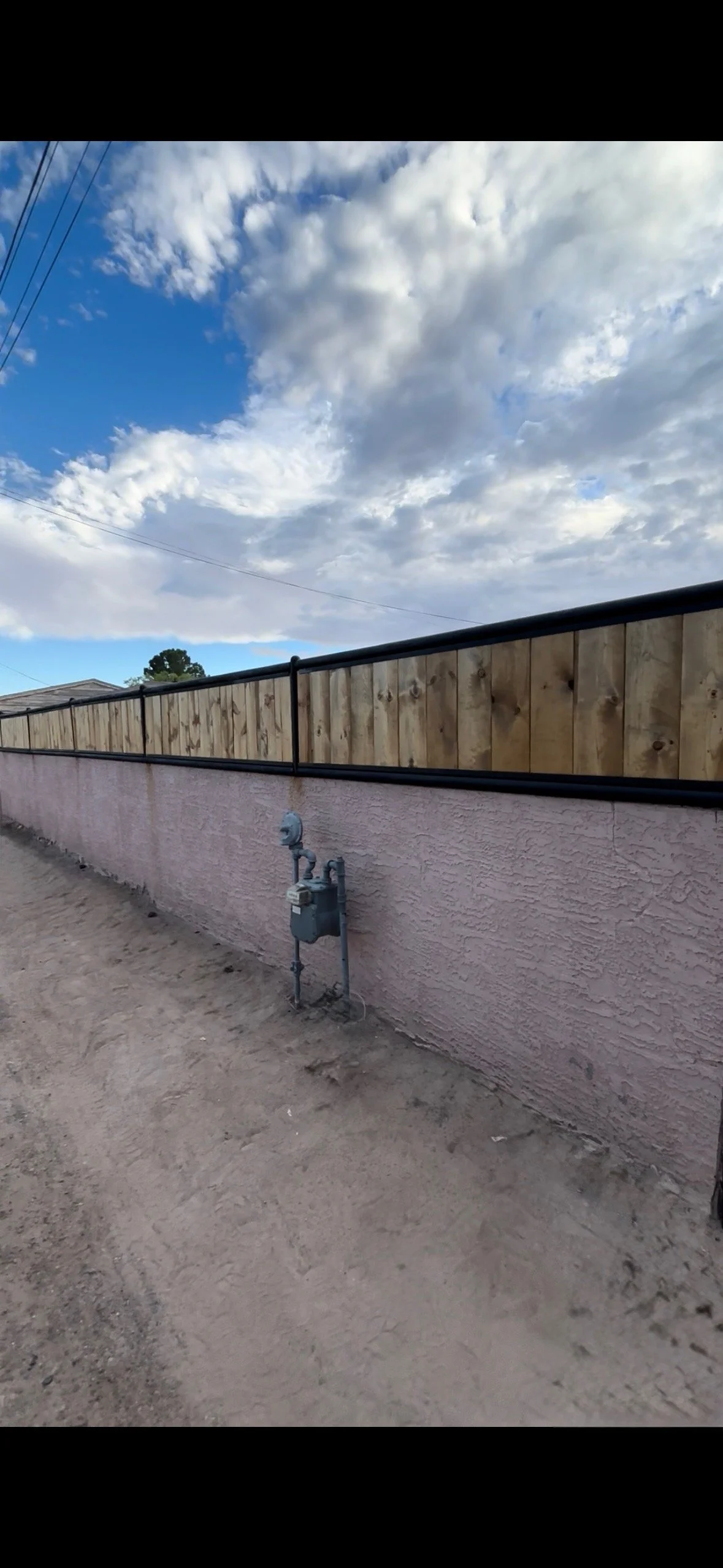 View of a pink stucco wall with a wooden fence on top, an outdoor gas meter attached to the wall, and a dirt area in the foreground. The sky above is partly cloudy with patches of blue.