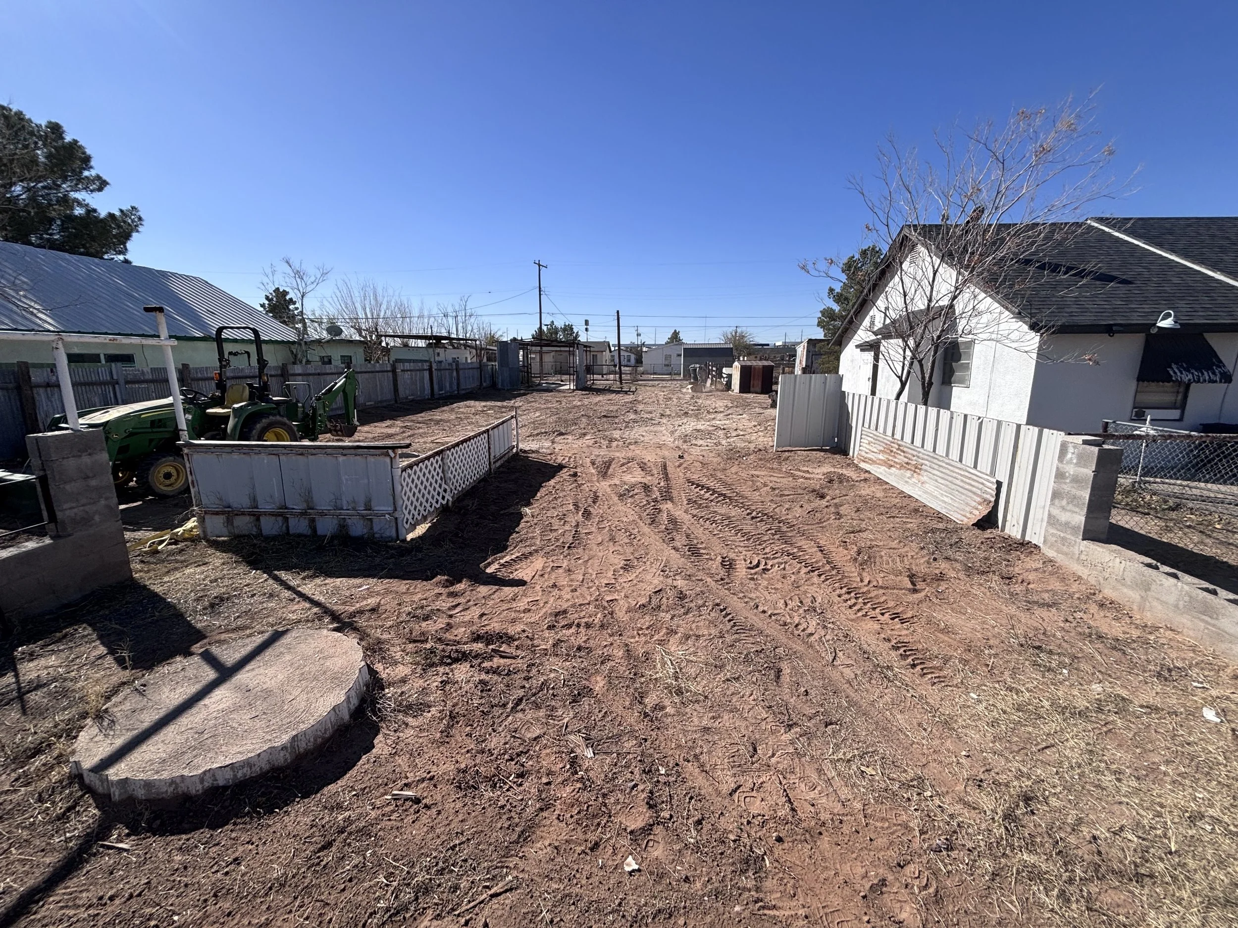 Empty backyard with dirt ground, some scattered debris, and a small fenced-in area with a green tractor. There are neighboring houses, fences, and leafless trees under a clear blue sky.