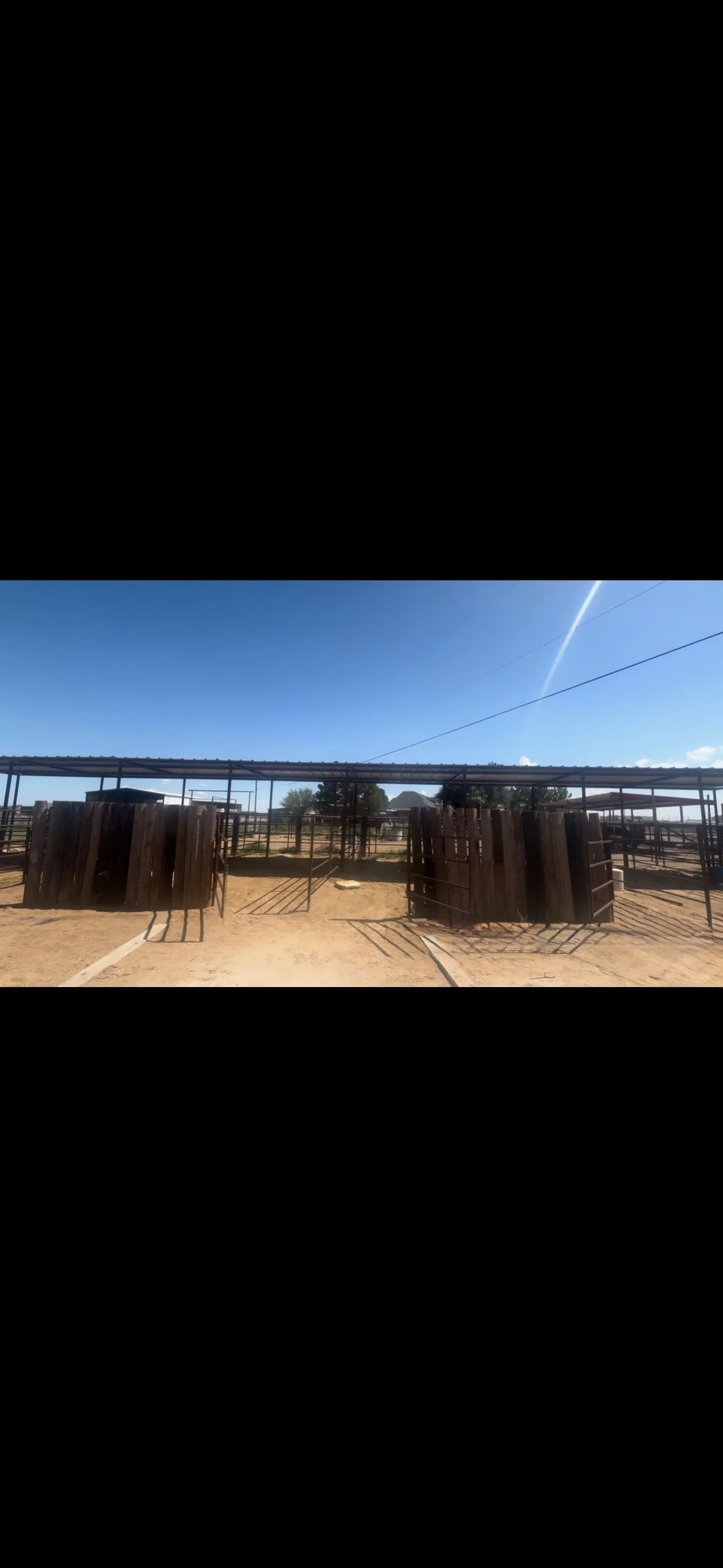 Outdoor livestock pen with wooden and metal fencing, open roof, and dirt ground, under a clear blue sky.
