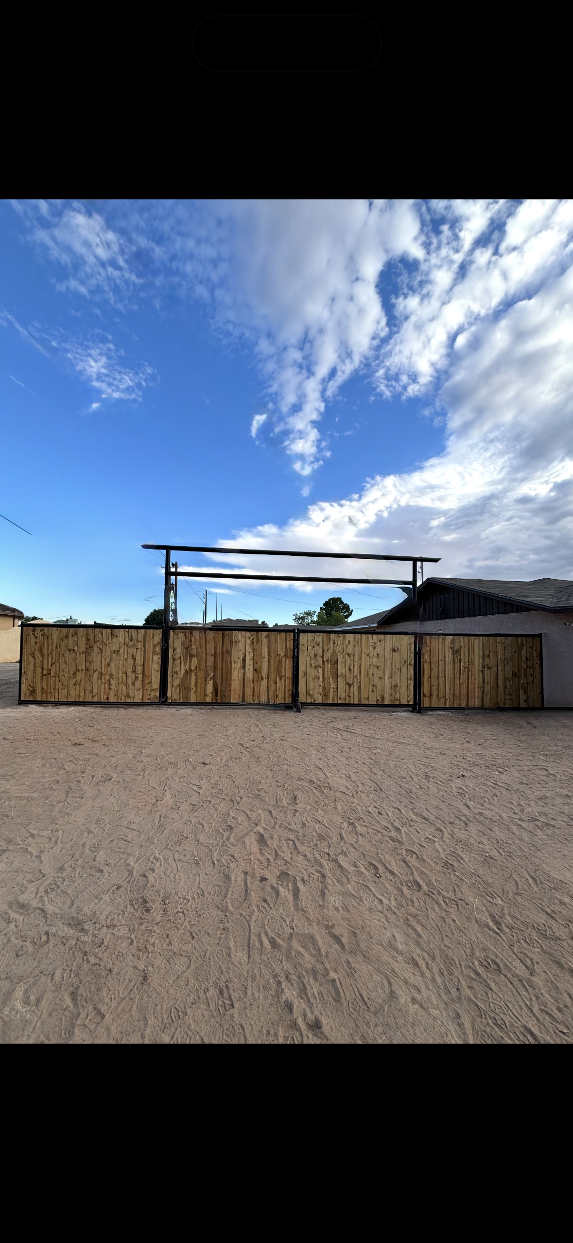 A wooden gate with a metal frame, set against a sandy ground and a partly cloudy blue sky.