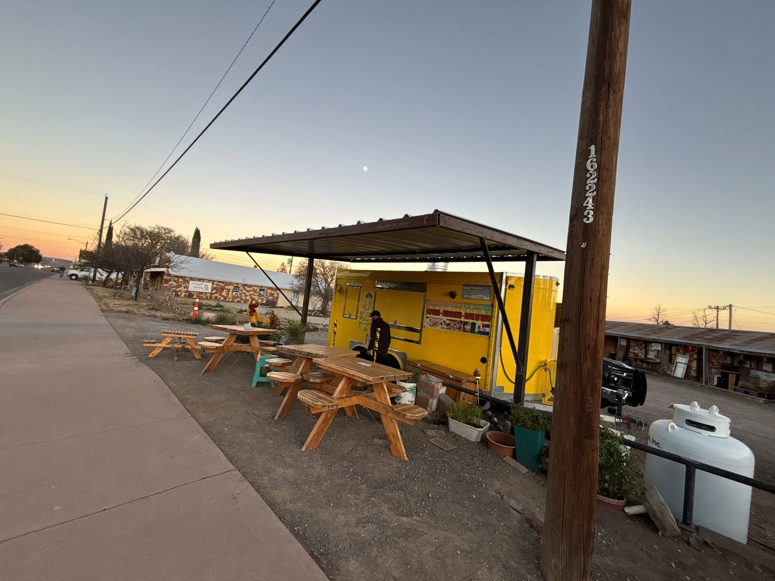 A food truck painted yellow with a menu, parked beside a sidewalk with wooden picnic tables, on a street during sunset with a partly visible moon and electric lines overhead.