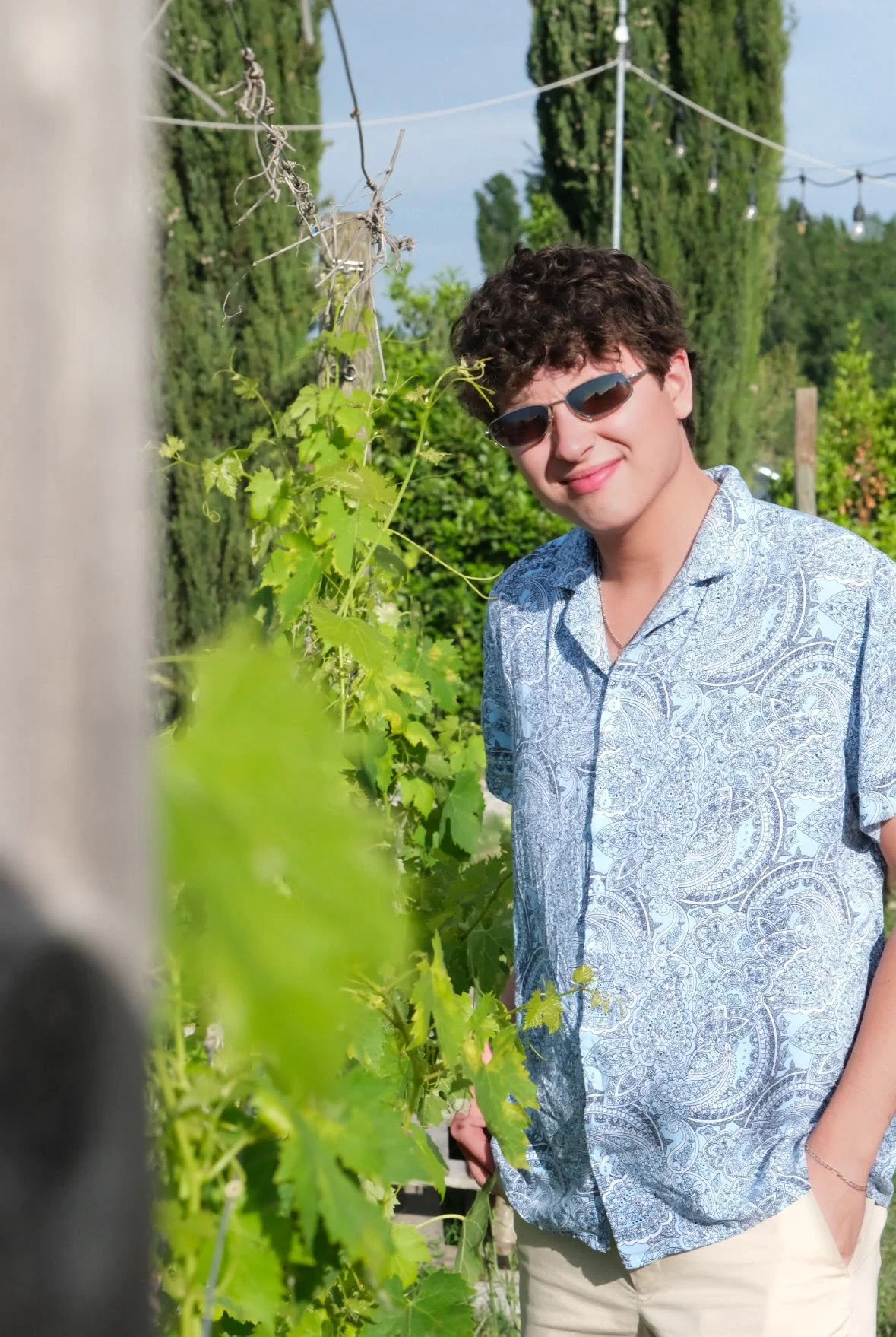 A young man wearing sunglasses and a patterned button-up shirt outdoors among green foliage, smiling at the camera.