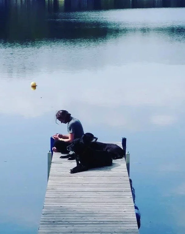 A girl and a black dog sitting on a wooden dock by a calm lake.