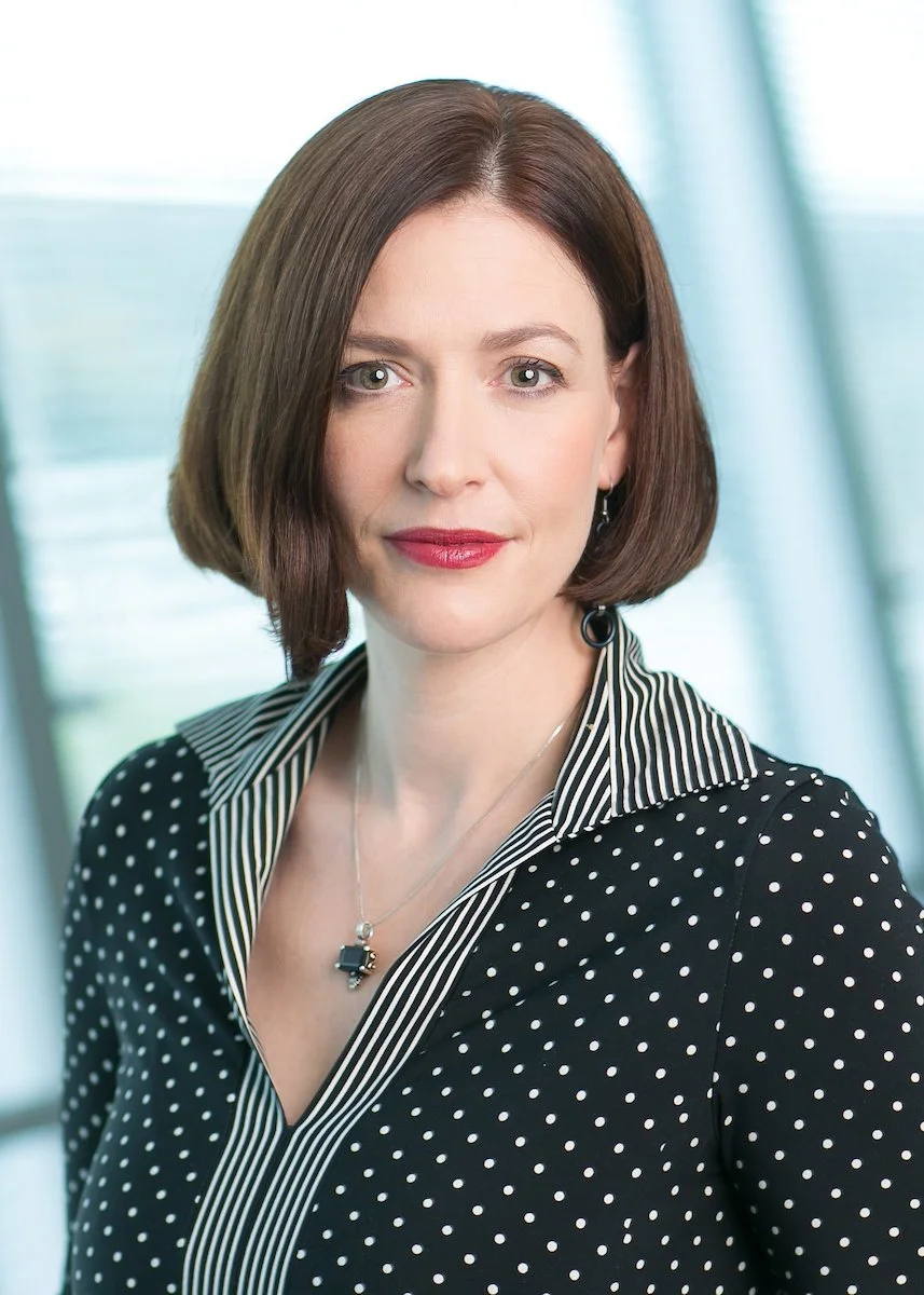 A woman with shoulder-length brown hair, green eyes, and red lipstick, wearing a black polka dot blouse with a striped collar, standing in front of a blurred window background.