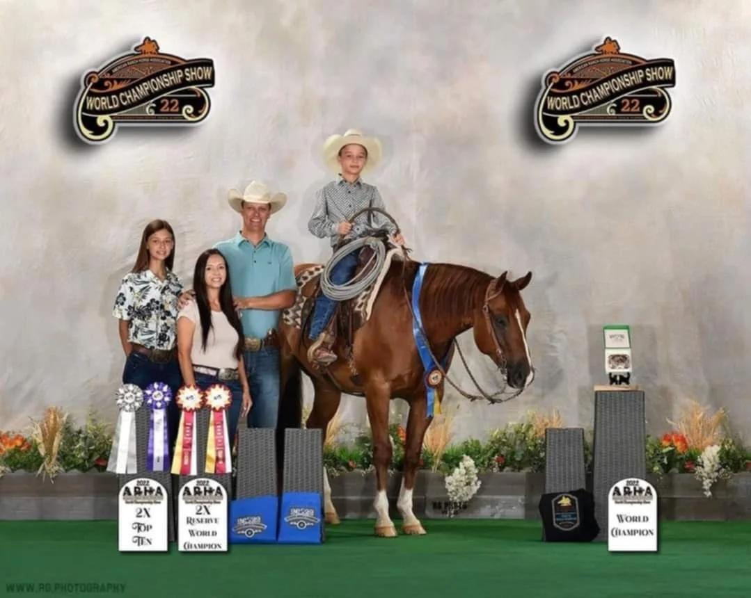 A horse rider with a cowboy hat, sitting on a brown horse with a blue ribbon, at an awards ceremony for the World Championship Show 2022. Three people stand beside the horse and rider, and ribbons and trophies are displayed on the floor.