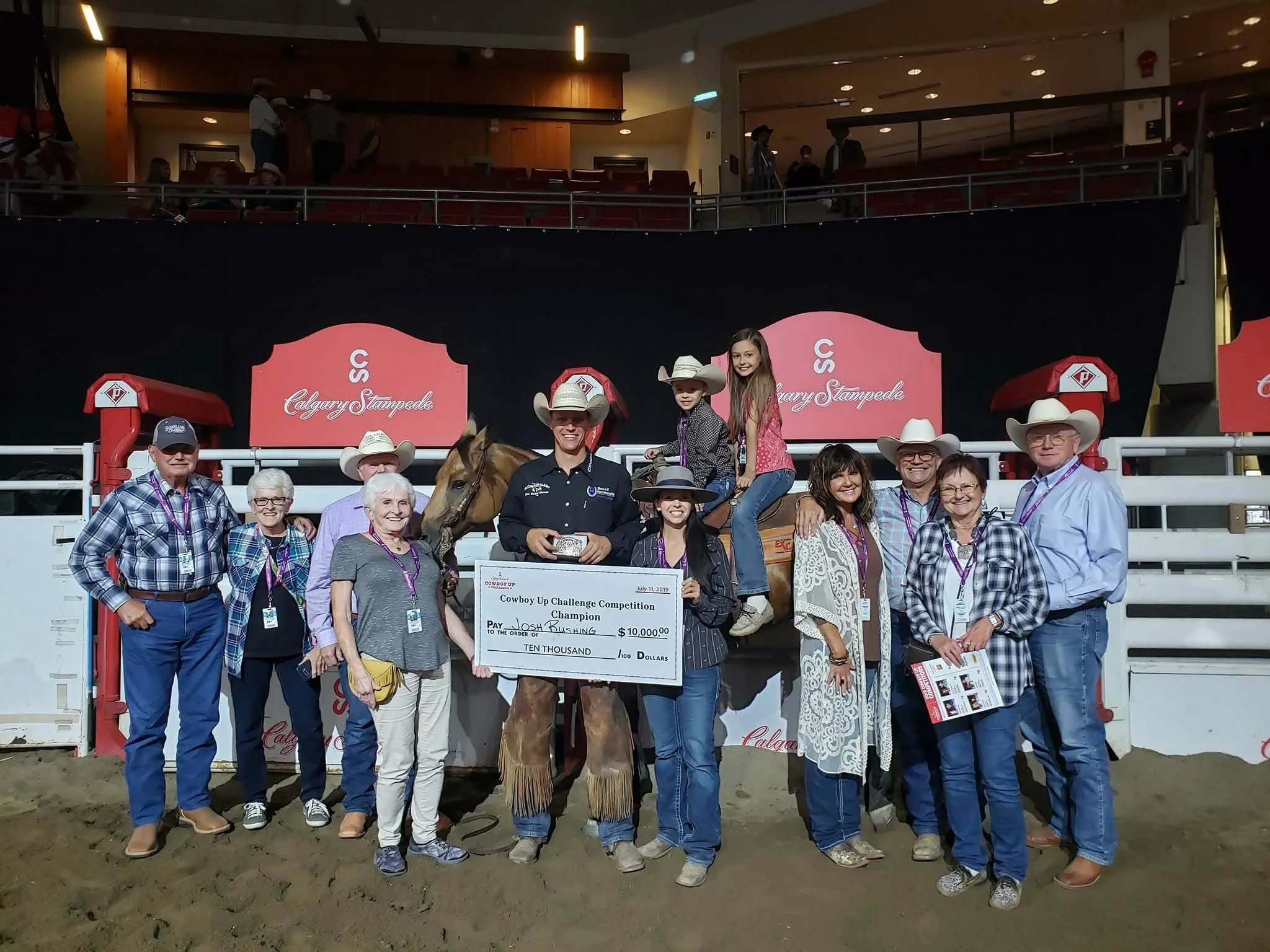 Group of people celebrating with a horse at the Calgary Stampede, holding a large championship check for $10,000, with some wearing cowboy hats and western attire.