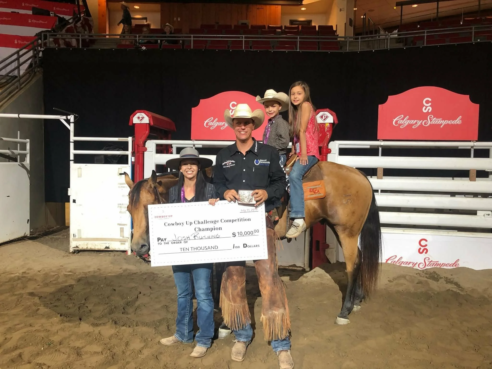 A group of four people, including two children, on horseback at an indoor rodeo arena. They are celebrating a victory with a large check for $10,000 awarded to Josh Rushing, with the words 'Cowboy Up Challenge Competition Champion.' The background sh