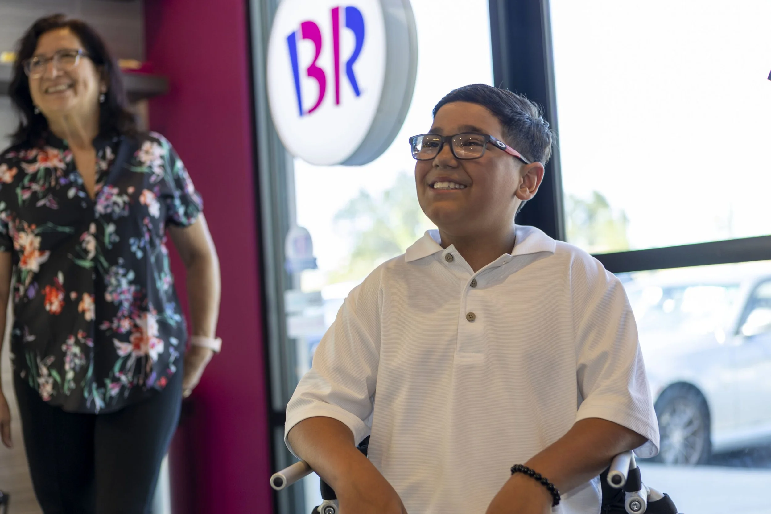 A young boy in a wheelchair wearing glasses and a white polo shirt, smiling inside a Baskin-Robbins restaurant with a woman standing nearby, also smiling, wearing a floral top. The restaurant's sign with 'BR' is visible in the background.