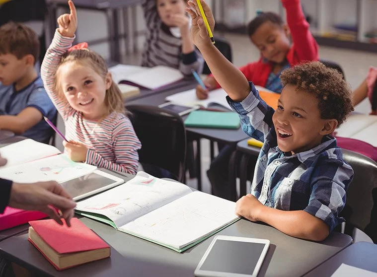 Young students in a classroom raising their hands and smiling during a lesson.