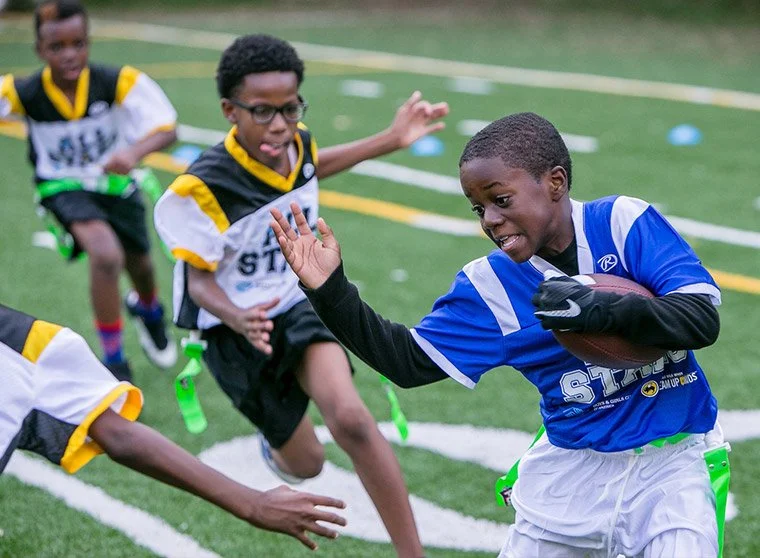 Children playing flag football on a field, with one boy in a blue jersey carrying a football, trying to avoid defenders.