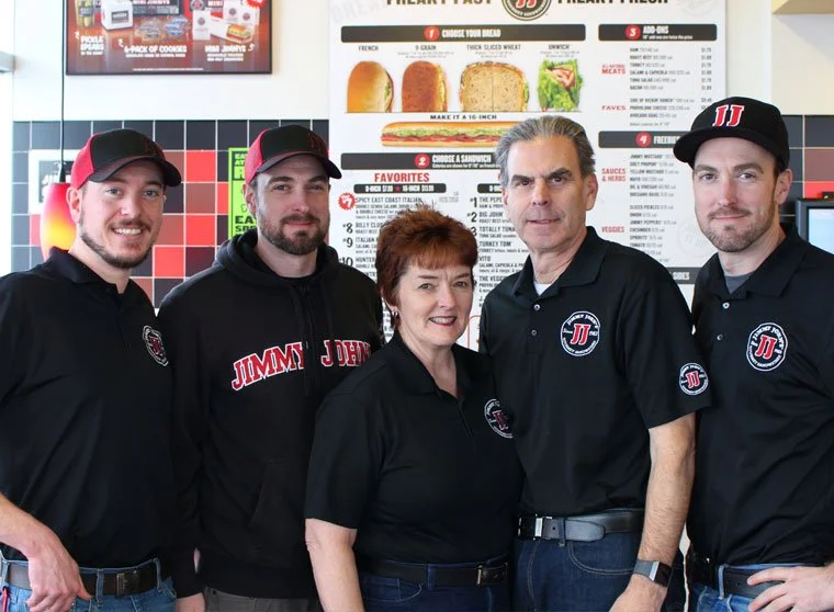 Five employees of Jimmy John's standing inside a sandwich shop, smiling at the camera with a menu board behind them.