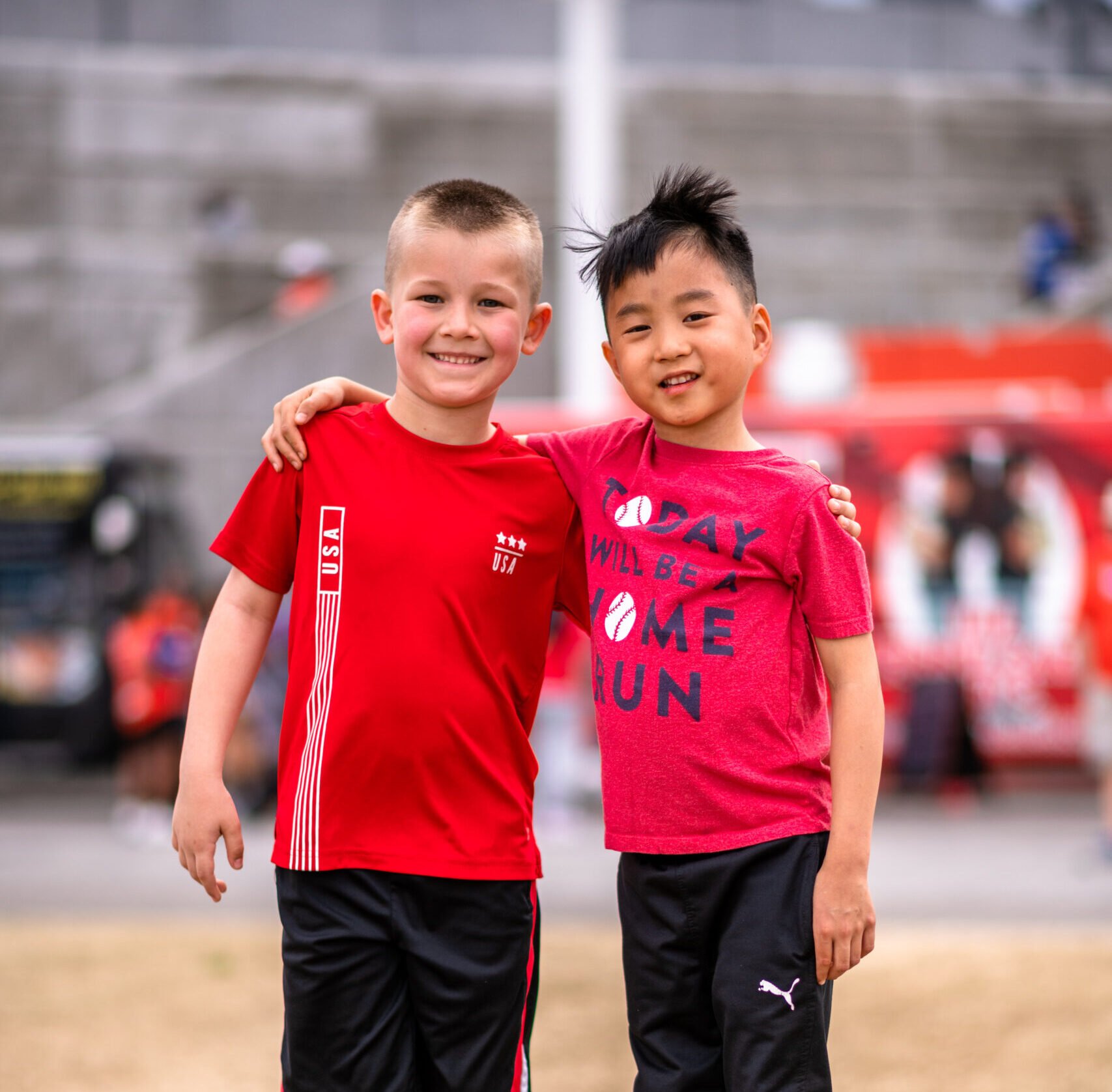 Two smiling boys wearing red shirts and black shorts standing arm in arm on a sports field, with bleachers and equipment in the background.