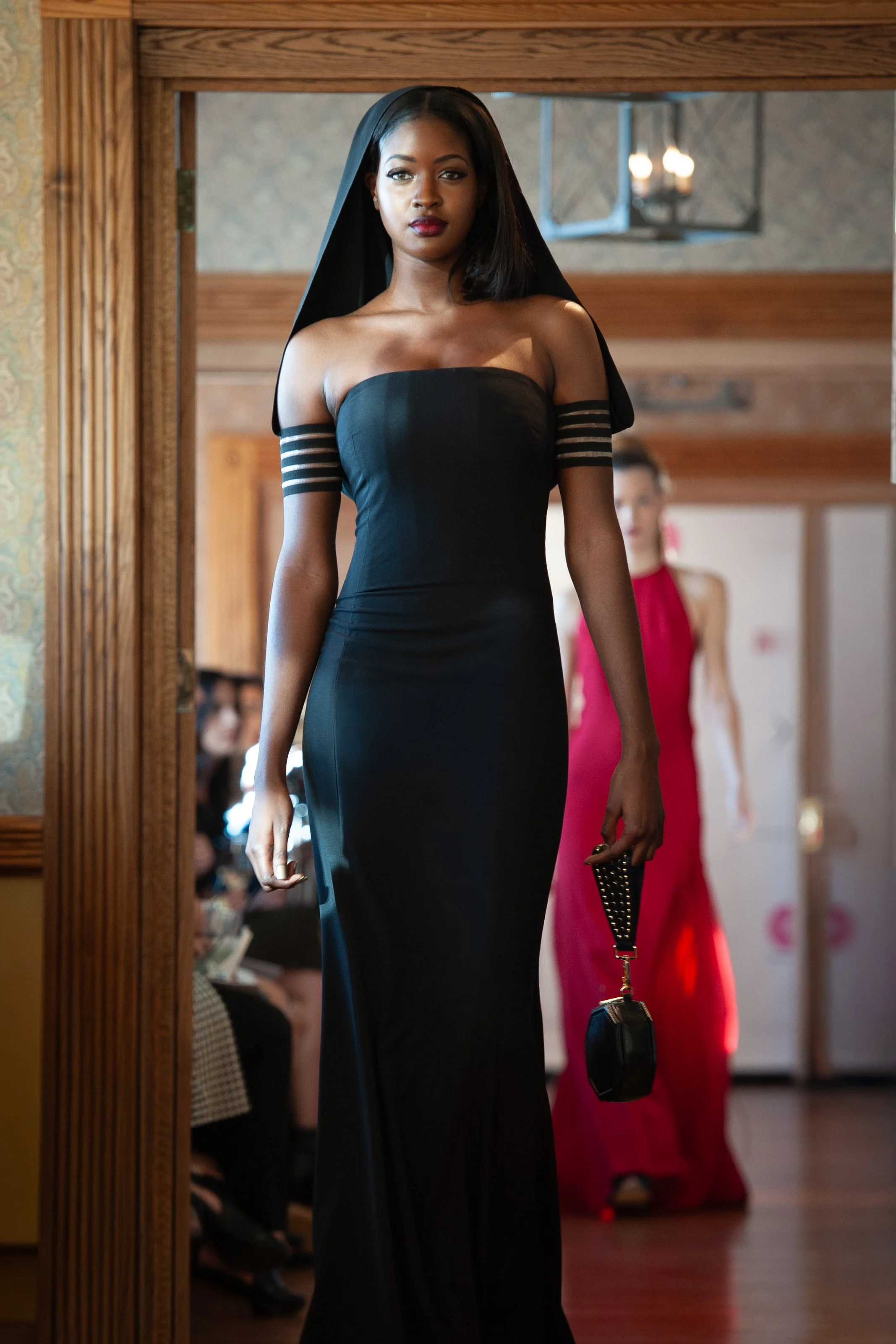 A woman in a black strapless evening gown, with sheer sleeve details, walking in a fashion show, holding a small handbag. There are other models and audience in the background.