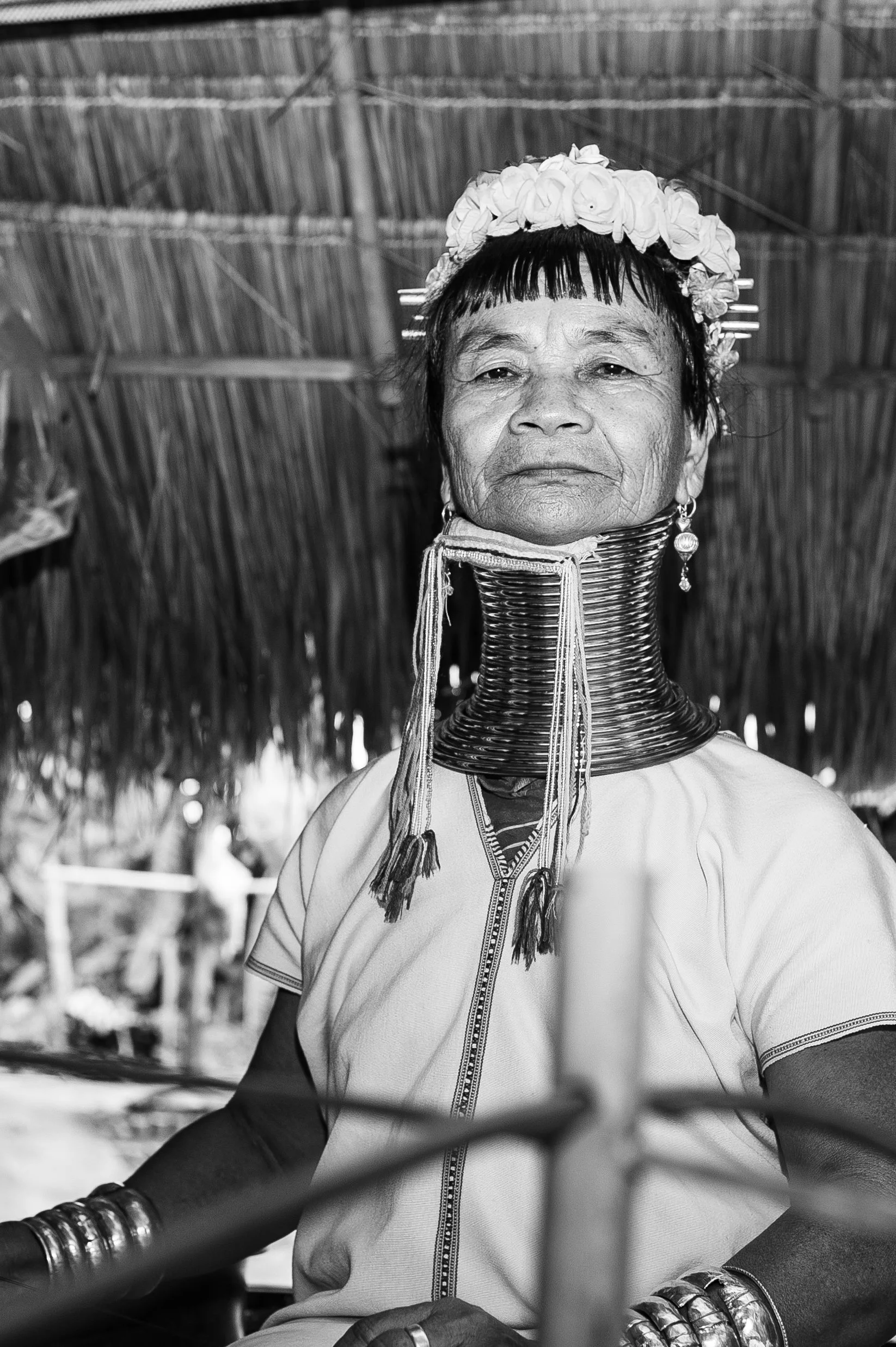 A woman from the Kayan tribe wearing traditional necklaces and clothing, sitting under a thatched roof.