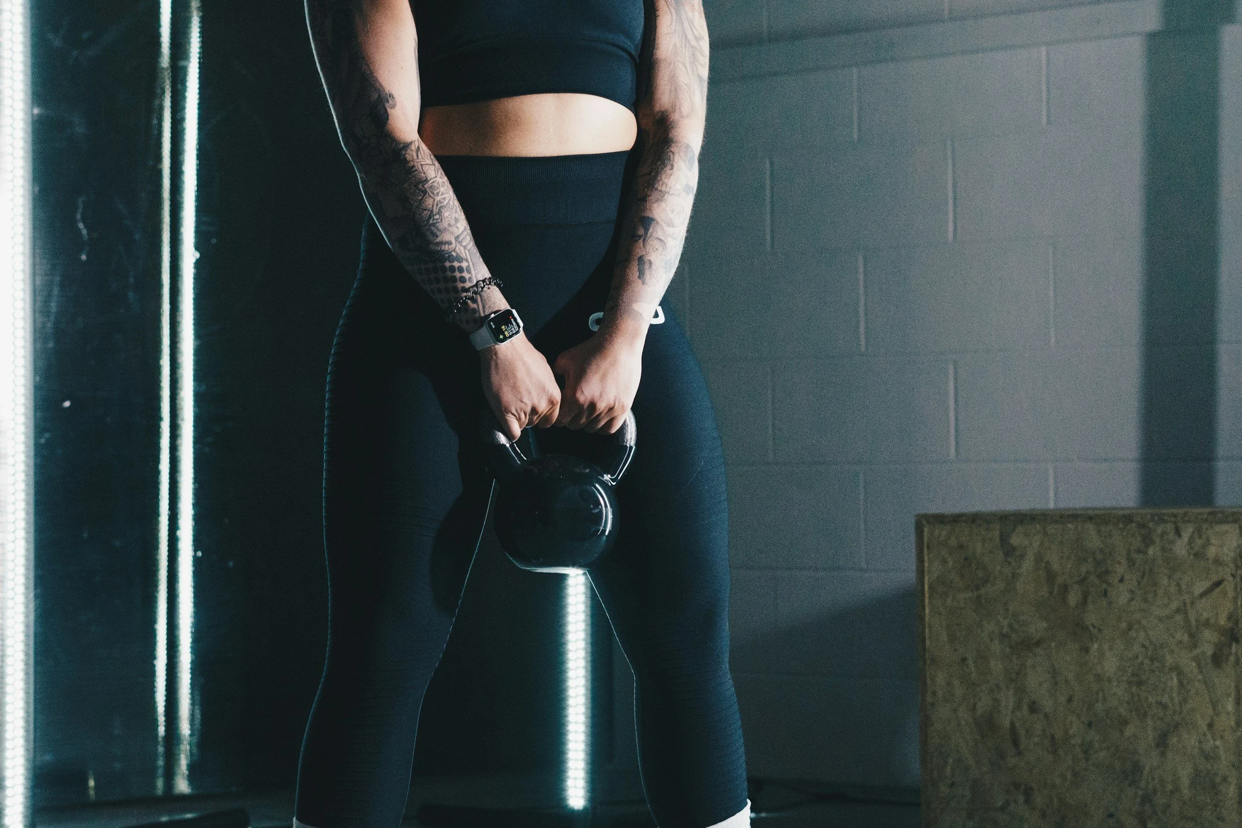 Person holding a black kettlebell in a gym, wearing black workout clothes, with tattoos on arms, smartwatch on wrist, against a gray wall and mirrored surface.