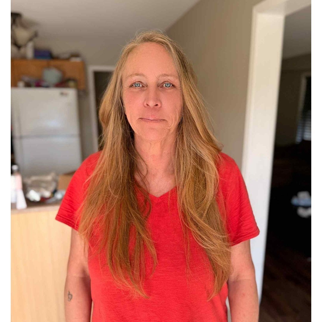 A woman with long, wavy, red hair wearing a red shirt standing indoors in a kitchen with a refrigerator and wooden cabinets in the background.