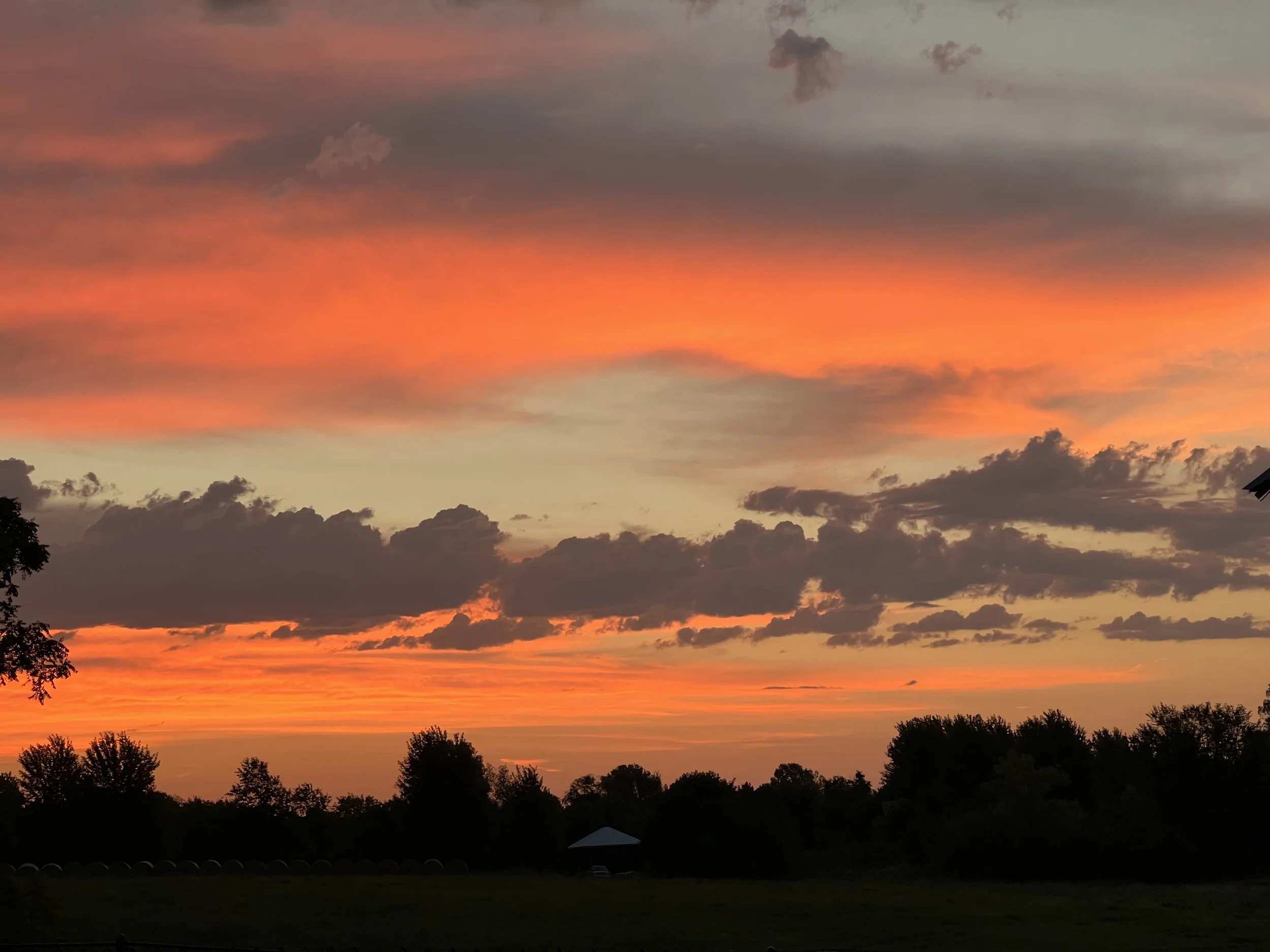 A colorful sunset sky with orange, pink, and purple hues, scattered clouds, and silhouettes of trees and a small building in the foreground.