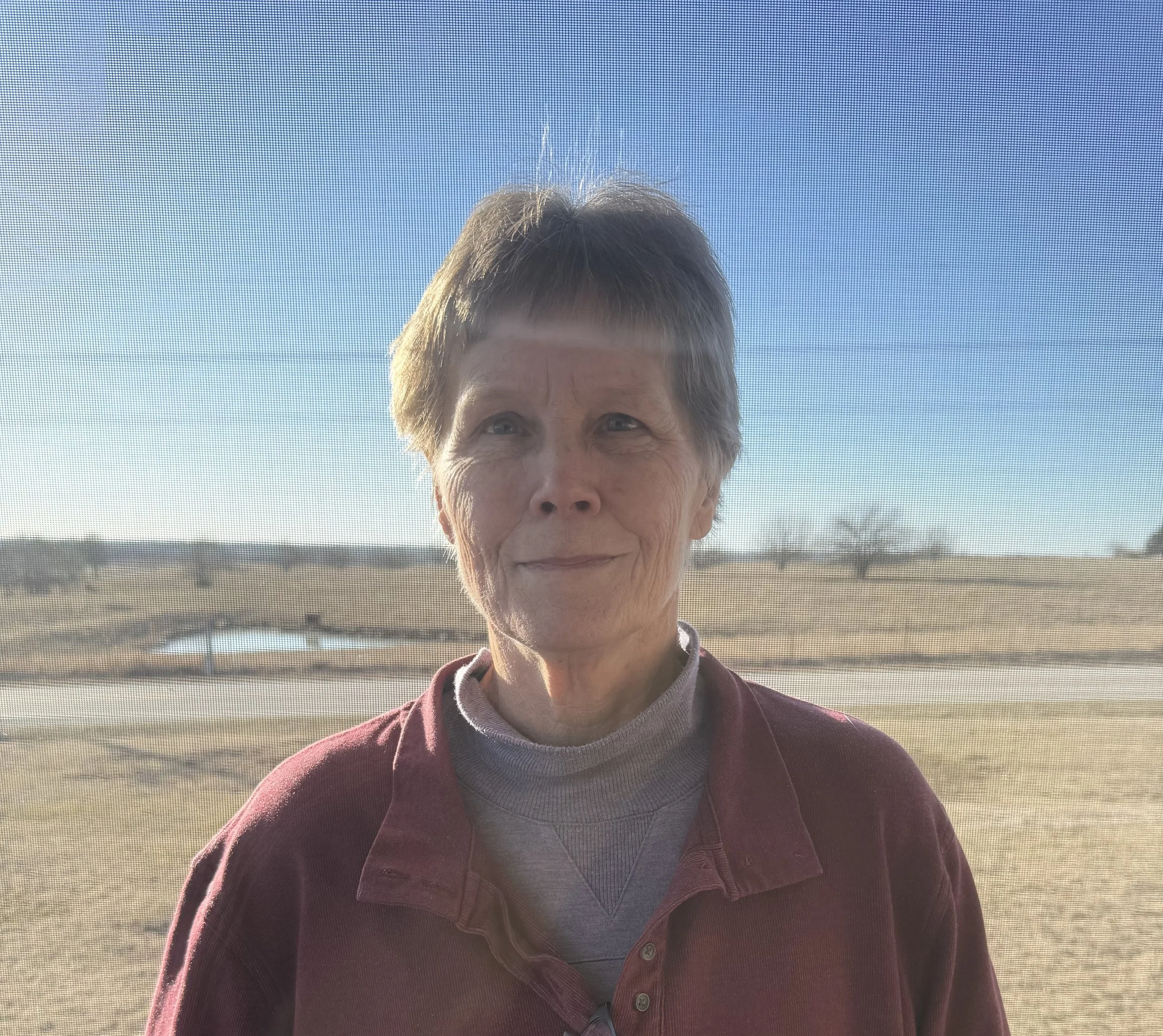 Older woman with short gray hair standing outside near a mesh screen, with a rural landscape of fields and a small pond in the background under a clear blue sky.