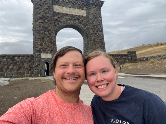 A smiling man and woman taking a selfie in front of a stone archway with cloudy sky and a hill in the background.