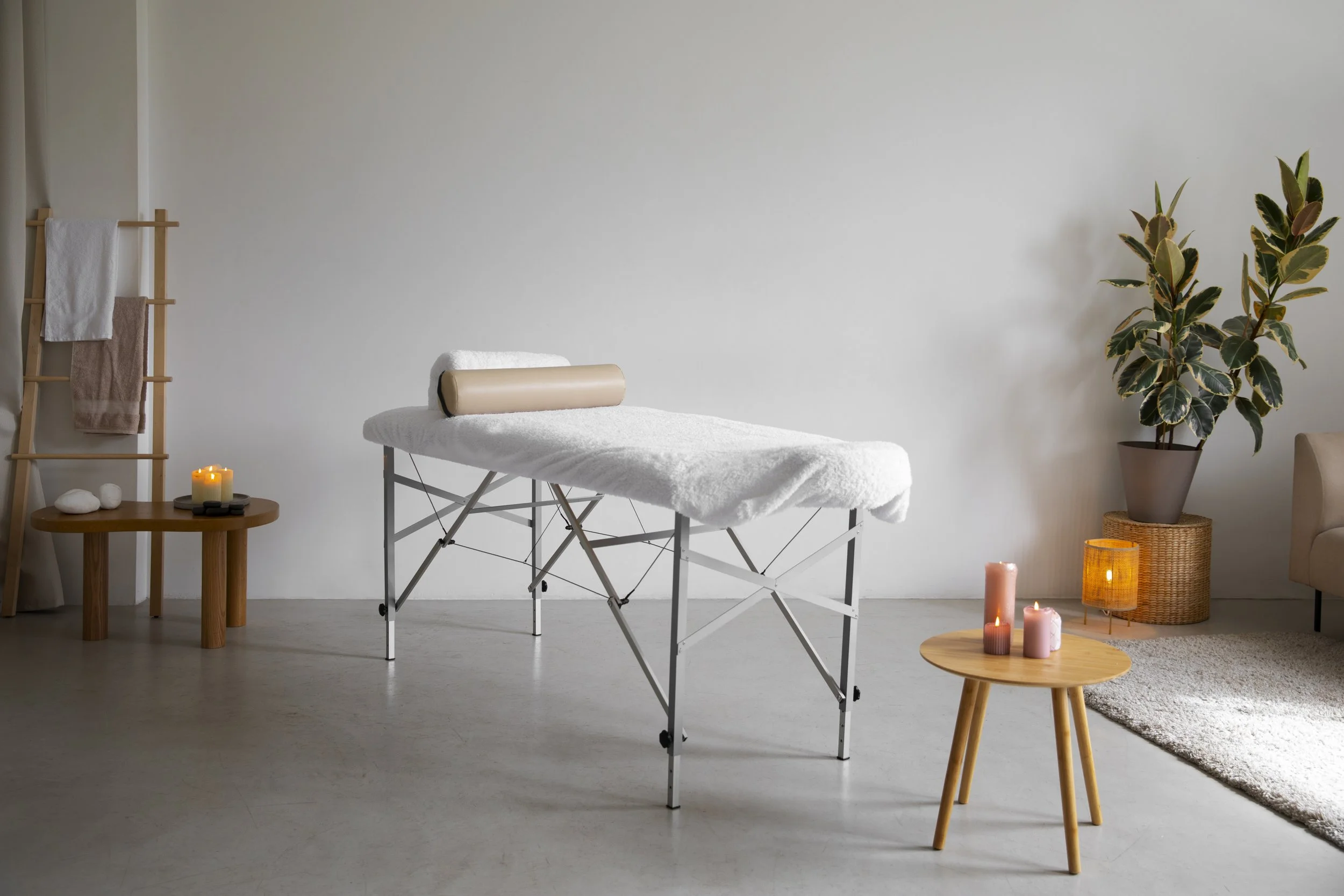 Spa or massage therapy room with a massage table covered in white towels, candles, a potted plant, and neutral-colored furniture and decor.
