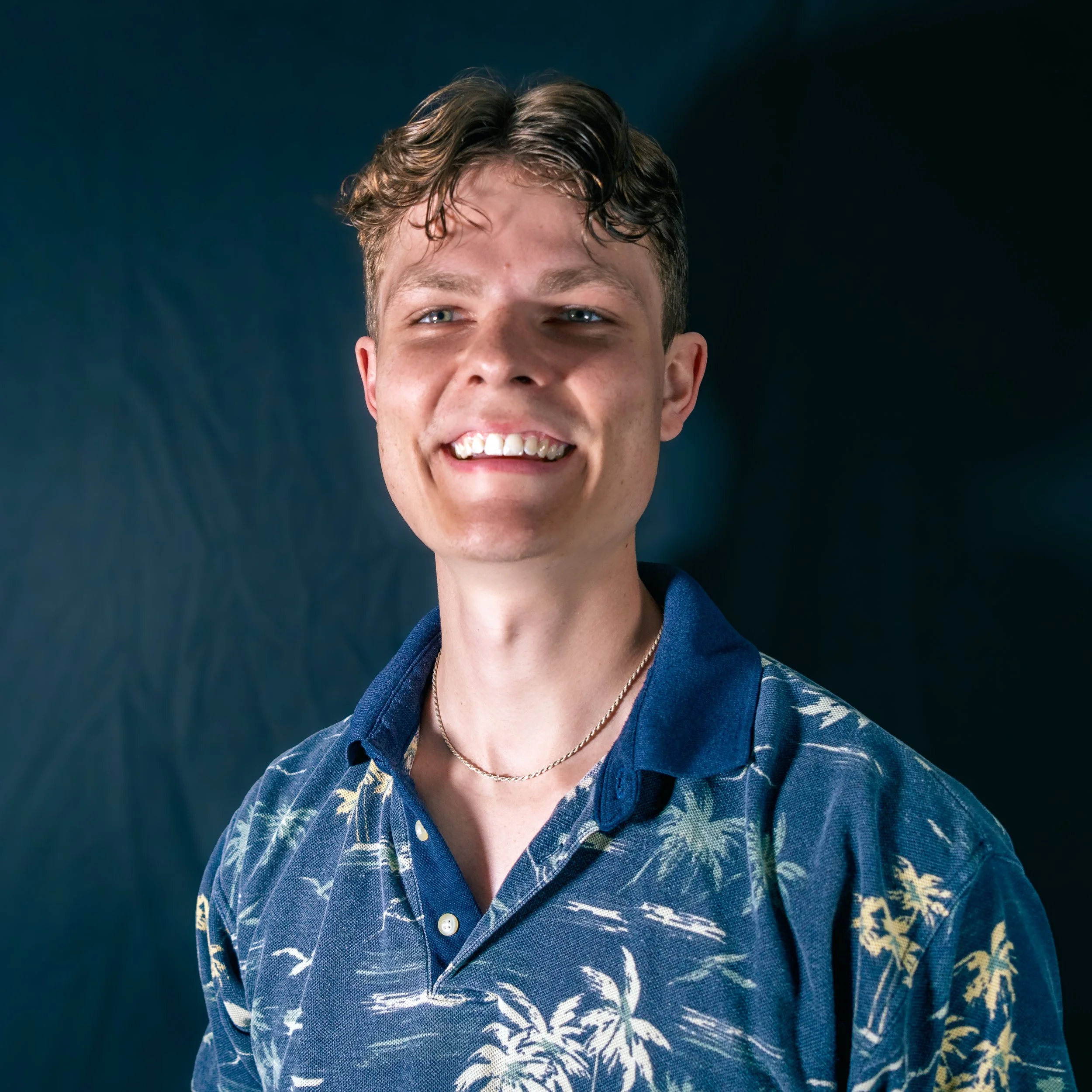 A smiling young man with wavy light brown hair, wearing a blue floral patterned short-sleeve button-up shirt and a thin gold necklace, standing against a dark background.