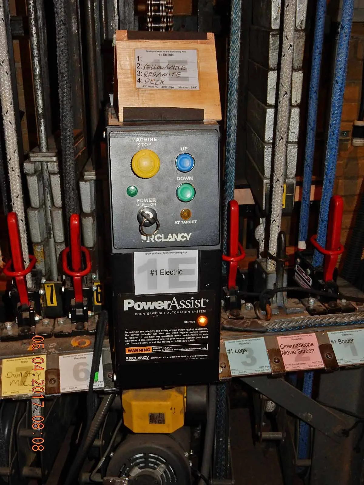 An electric control board for stage equipment with buttons labeled UP, DOWN, STOP, and AT TARGET. There are color-coded labels and a speed control switch. Several cables and clamps are attached, with a small notepad on top listing color codes for wiring. The control board is mounted above a motor and surrounded by various cables and metal bars.