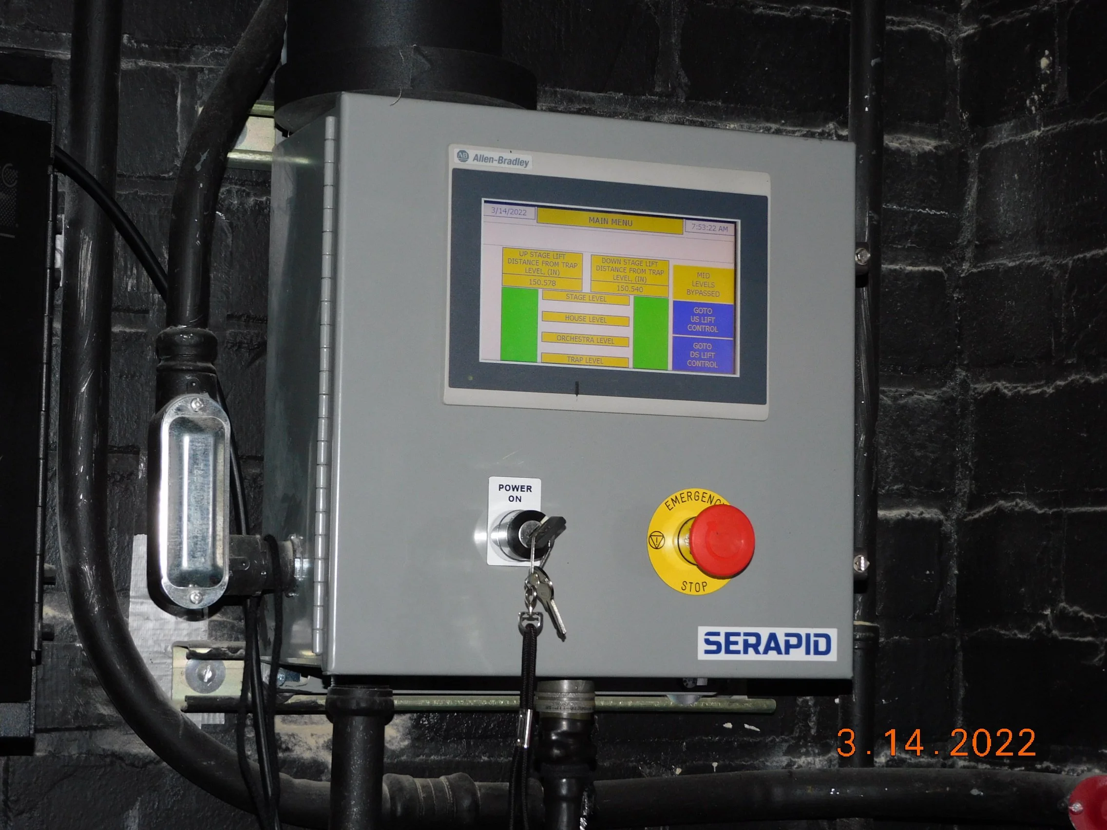 Industrial control panel with a touchscreen display, emergency stop button, power switch, and associated wiring mounted on a black brick wall.