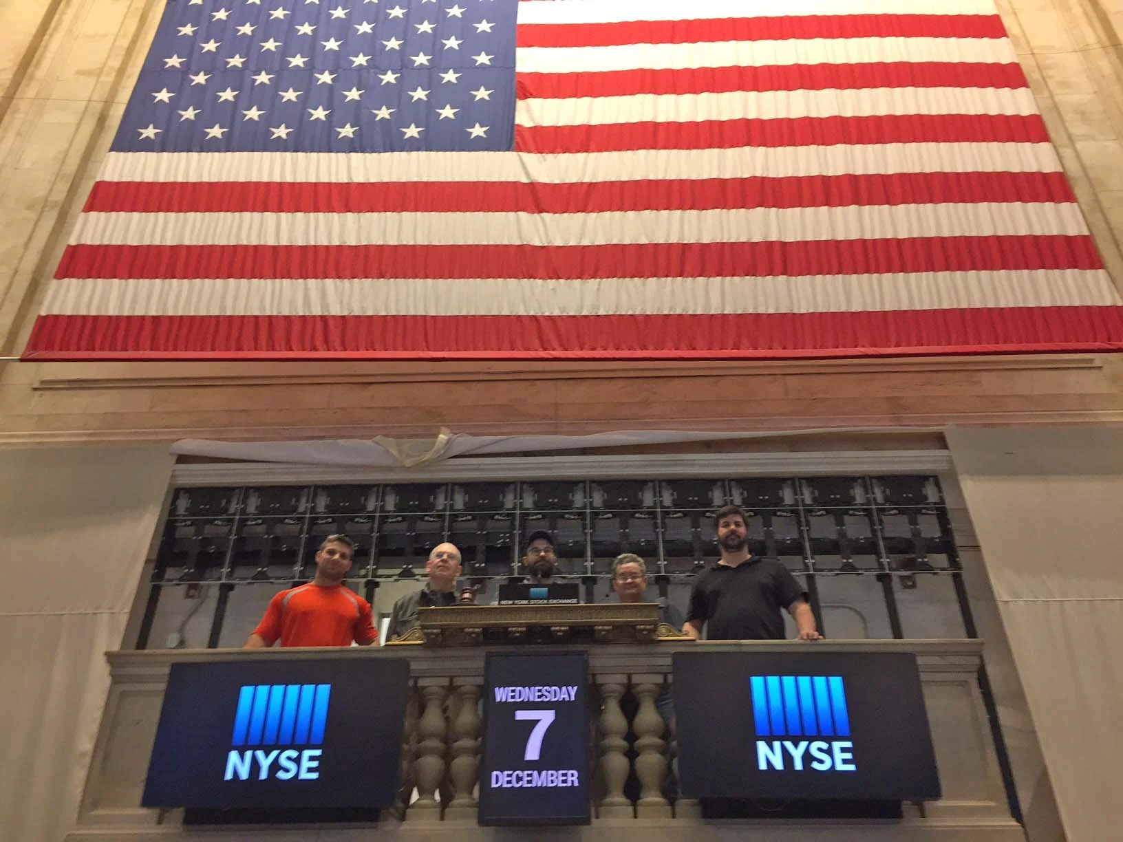 Large American flag hanging above a stock trading floor with four people standing at a trading post. Digital boards display NYSE and date December 7, Wednesday.