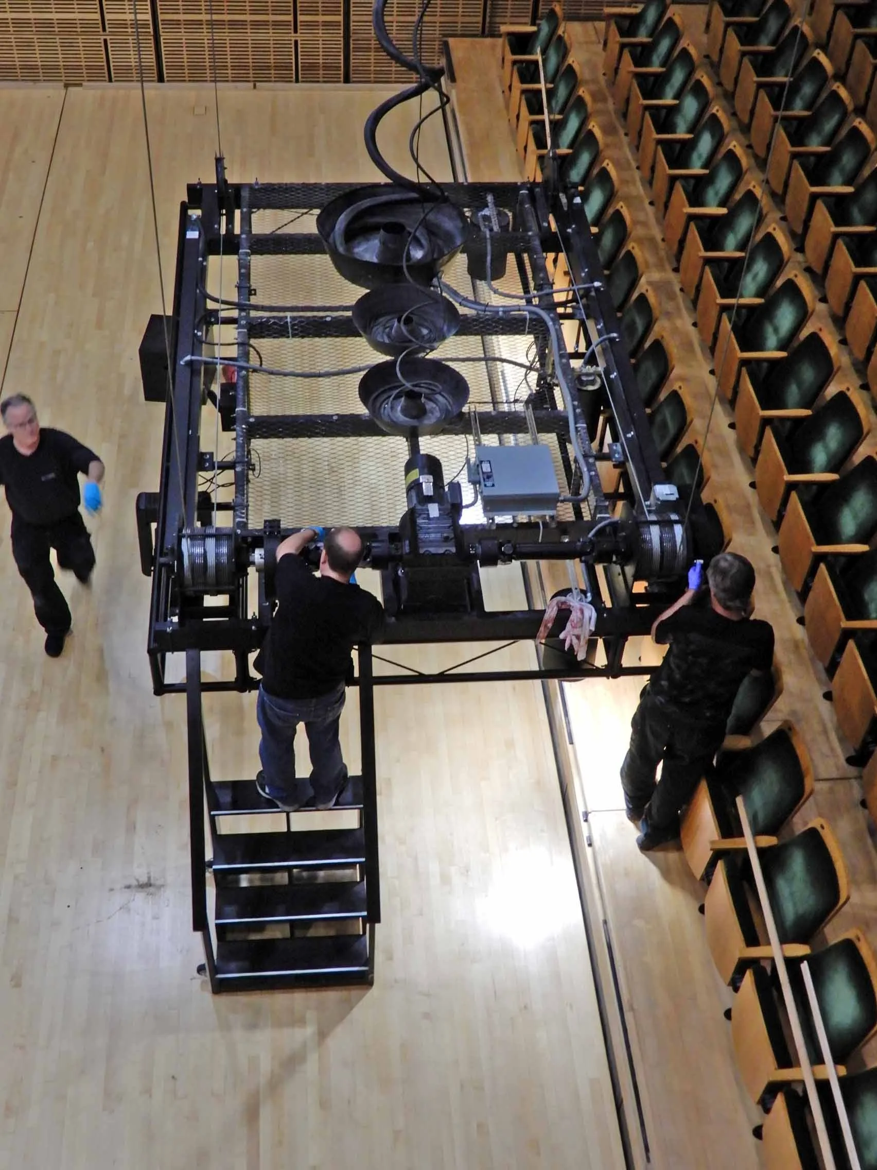 Three people working on a large mechanical stage apparatus in a room with wooden floors and tiered seating.