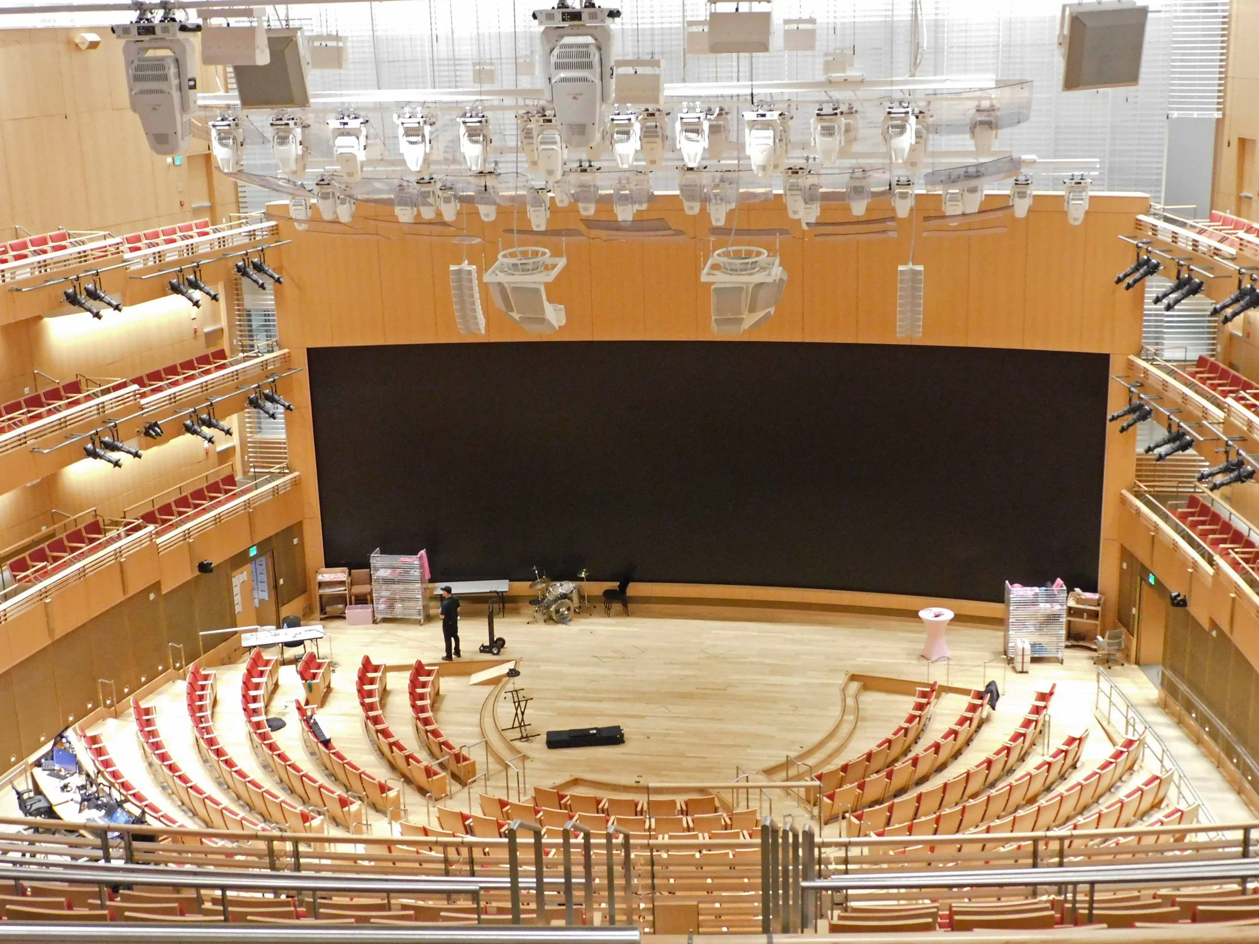 Large venue from back towards stage, with lots of rigging, lighting and acoustic panels. Large LED screen at back of stage.