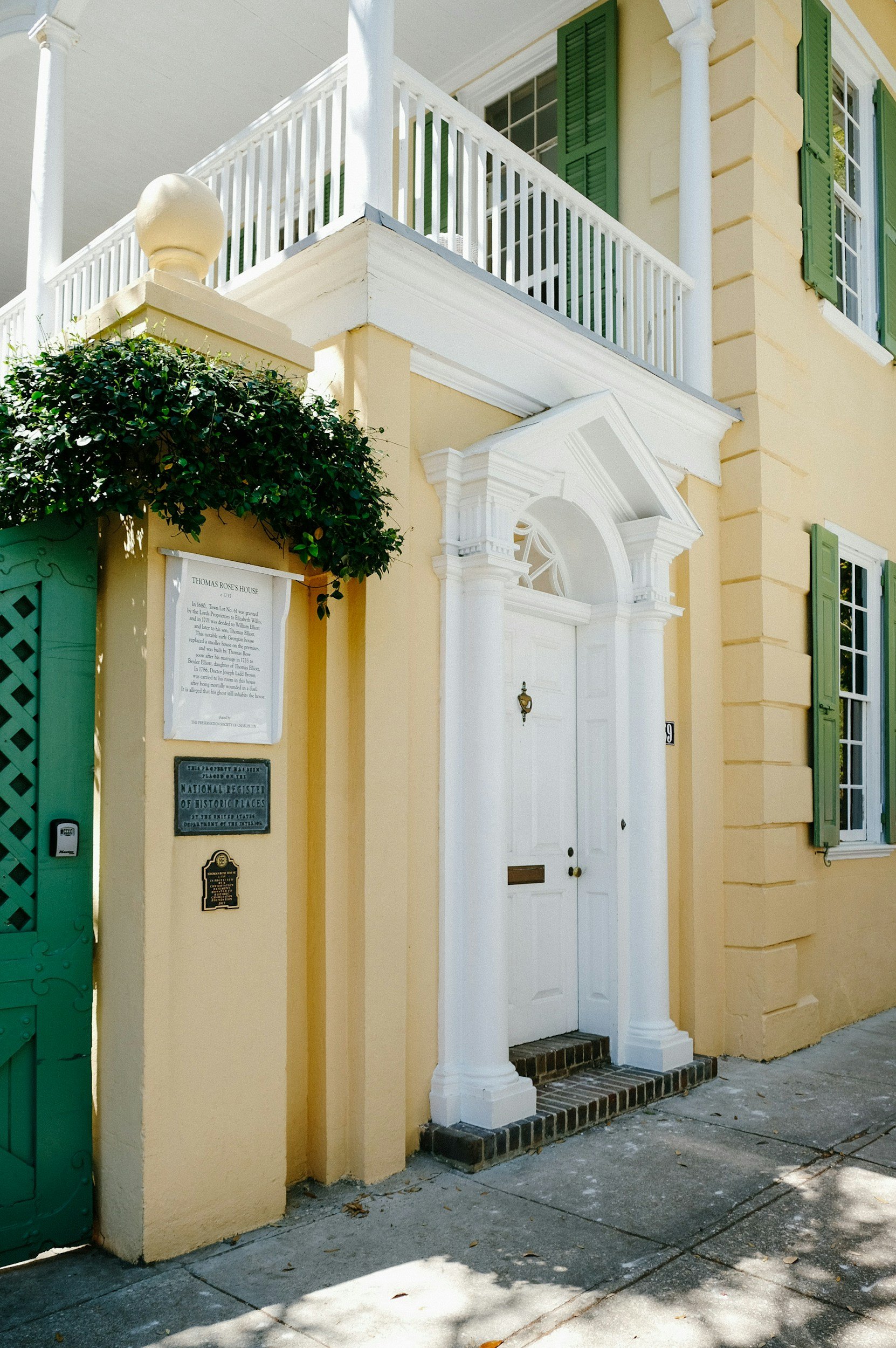 Yellow historic house with white door flanked by white columns, green shutters, and plaques on the wall, with a sidewalk in front.