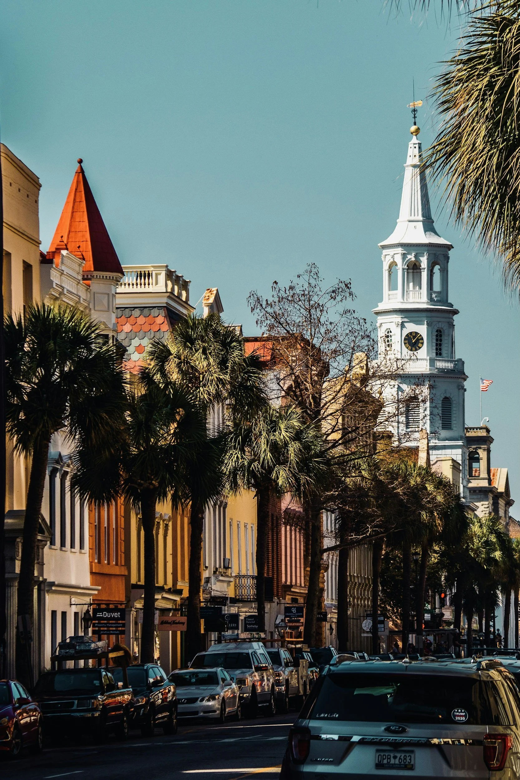 Street view with colorful buildings, palm trees, parked cars, and a white clock tower with a weather vane at the top.
