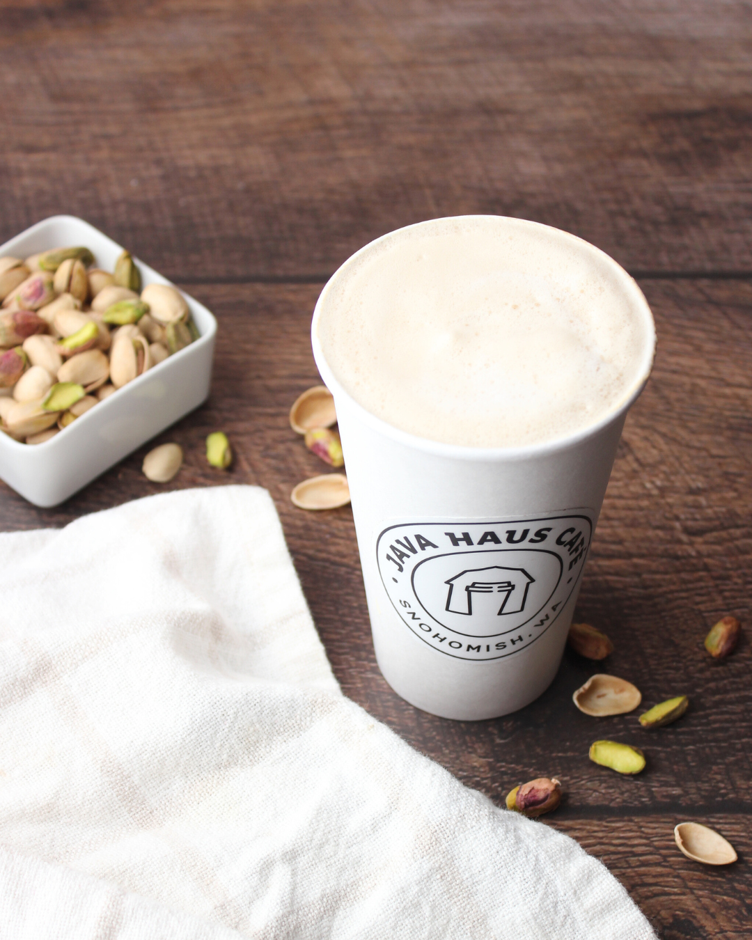 A white paper cup of coffee with foam on top, sitting on a wooden table with pistachio nuts and shells scattered around and a bowl of pistachios beside it.