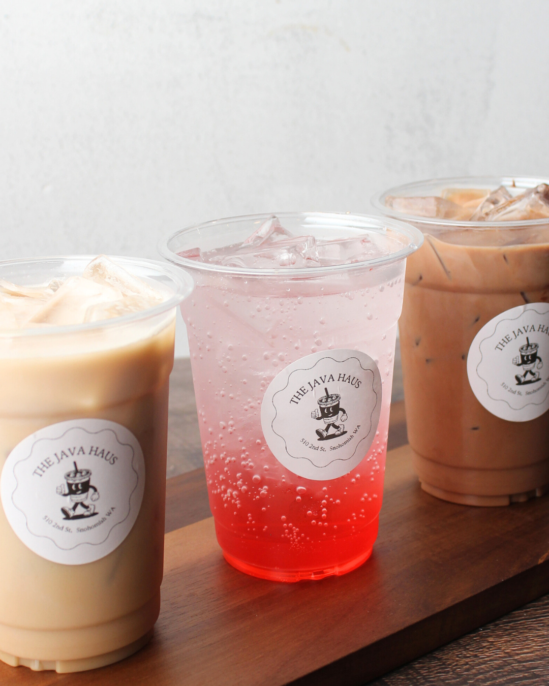 Three iced beverages with different colors on a wooden tray, each in a clear plastic cup with a logo reading 'The Java Haus'.
