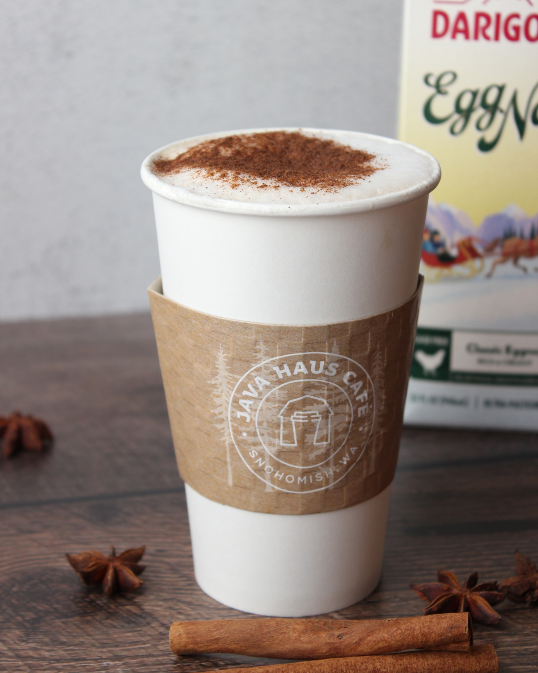 Hot beverage in white paper cup with a cardboard sleeve, topped with cocoa powder, placed on a wooden surface with cinnamon sticks and star anise, background shows a bag of Darigold Egg Nog.