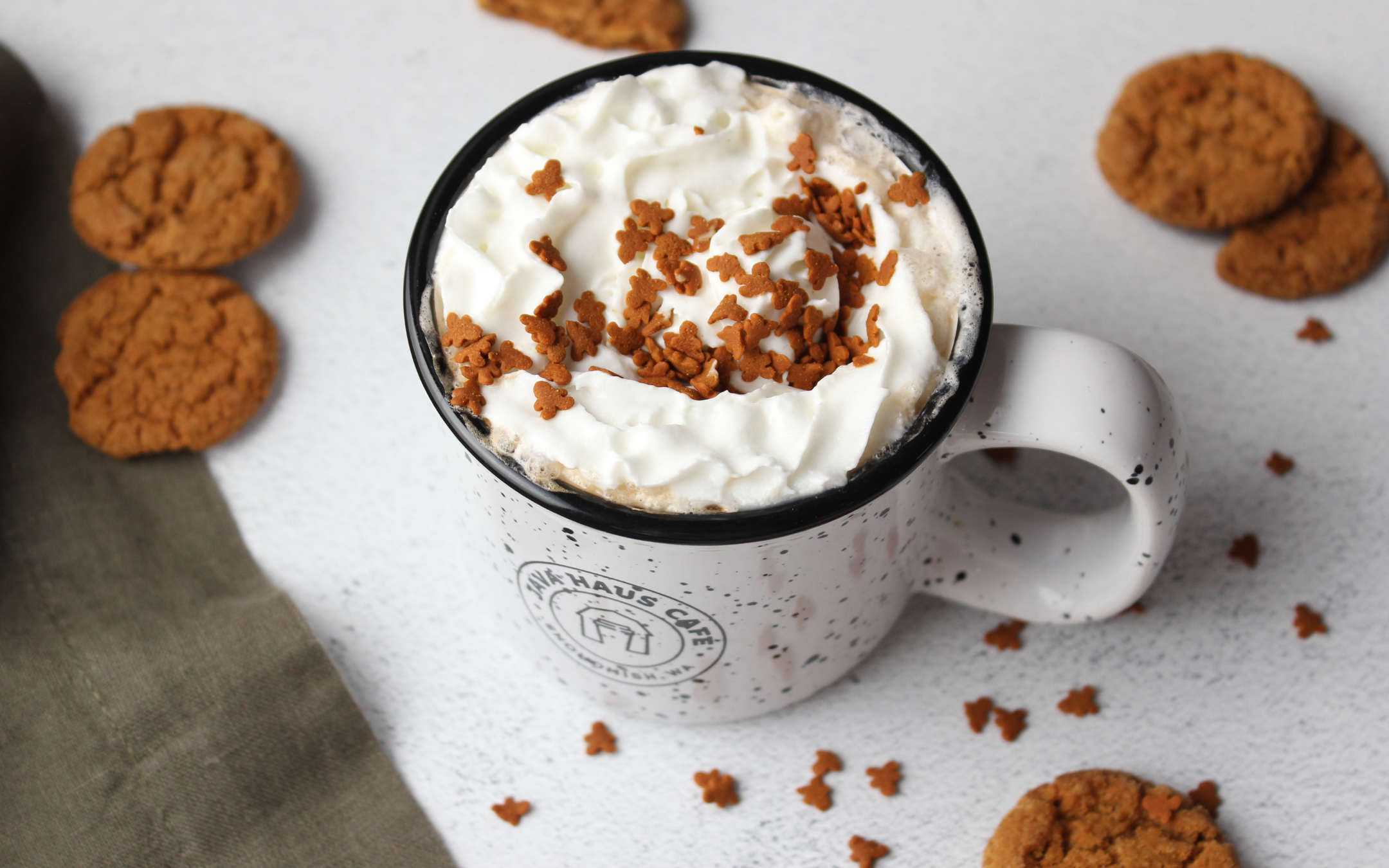 A white speckled mug filled with hot chocolate topped with whipped cream and chocolate sprinkles, surrounded by cookies on a white surface.