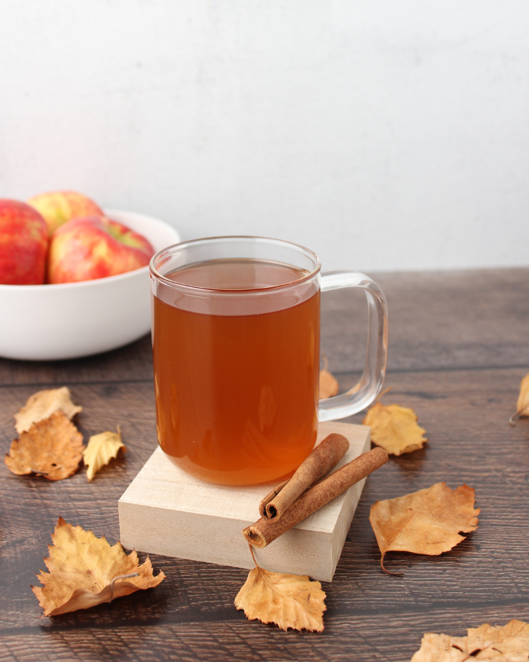 A glass mug of hot apple cider with two cinnamon sticks on a wooden table surrounded by fallen autumn leaves, with a white bowl of apples in the background.