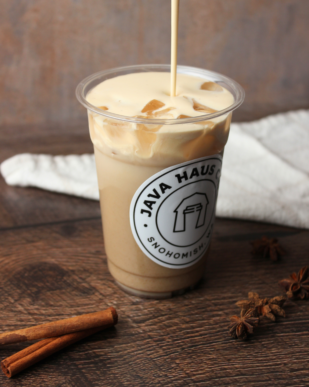 Iced coffee with cream in a plastic cup on a wooden surface, with cinnamon sticks, star anise, and a white cloth in the background.