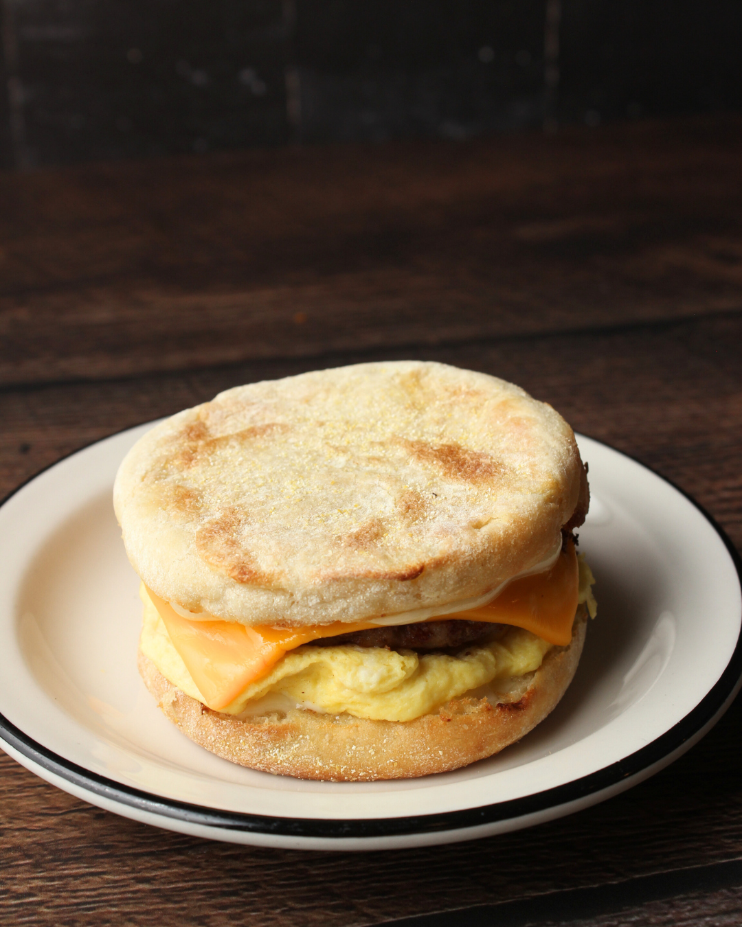 Breakfast sandwich with egg, cheddar cheese, sausage patty, on an English muffin, served on a white plate with a dark wood table background.