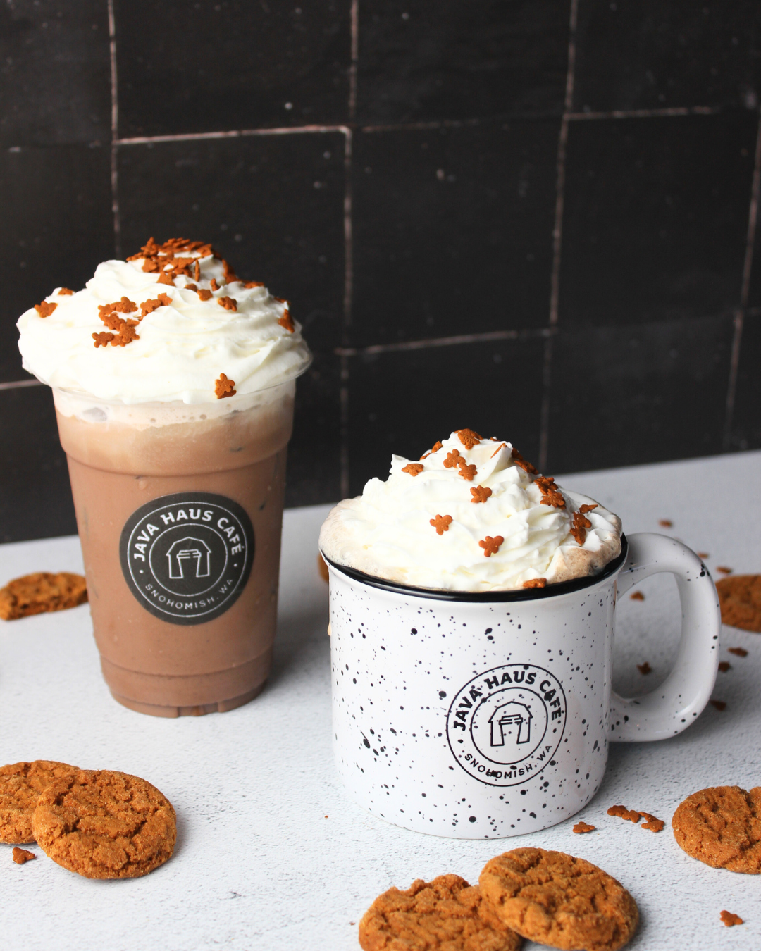 Two drinks topped with whipped cream and sprinkles, cookies scattered on a white surface, black and white speckled mugs, and a black tiled background.
