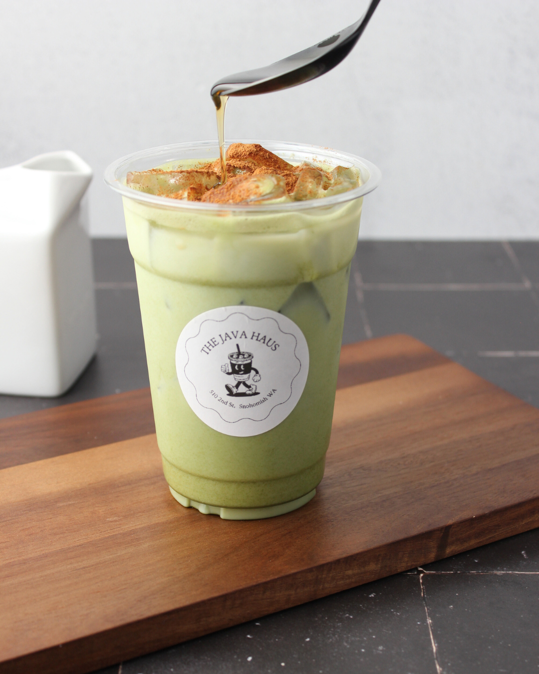 Iced matcha latte in a plastic cup with a label from The Java Haus, topped with whipped cream, cinnamon, and a drizzle of caramel or honey, on a wooden board.