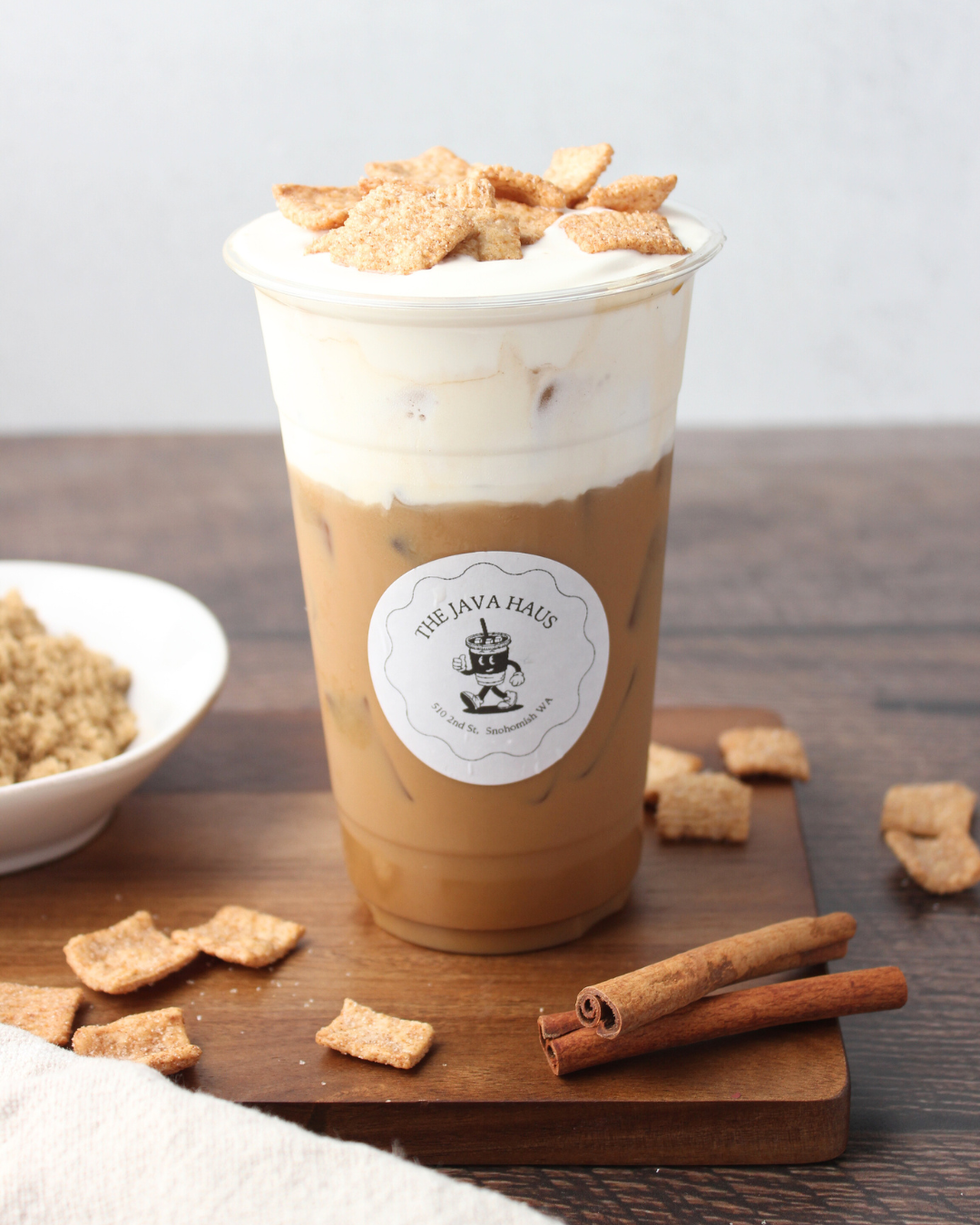 Iced coffee beverage topped with whipped cream and cinnamon toast crunch cereal in a plastic cup with a 'The Java Haus' logo, placed on a wooden board with cinnamon sticks and cereal around.