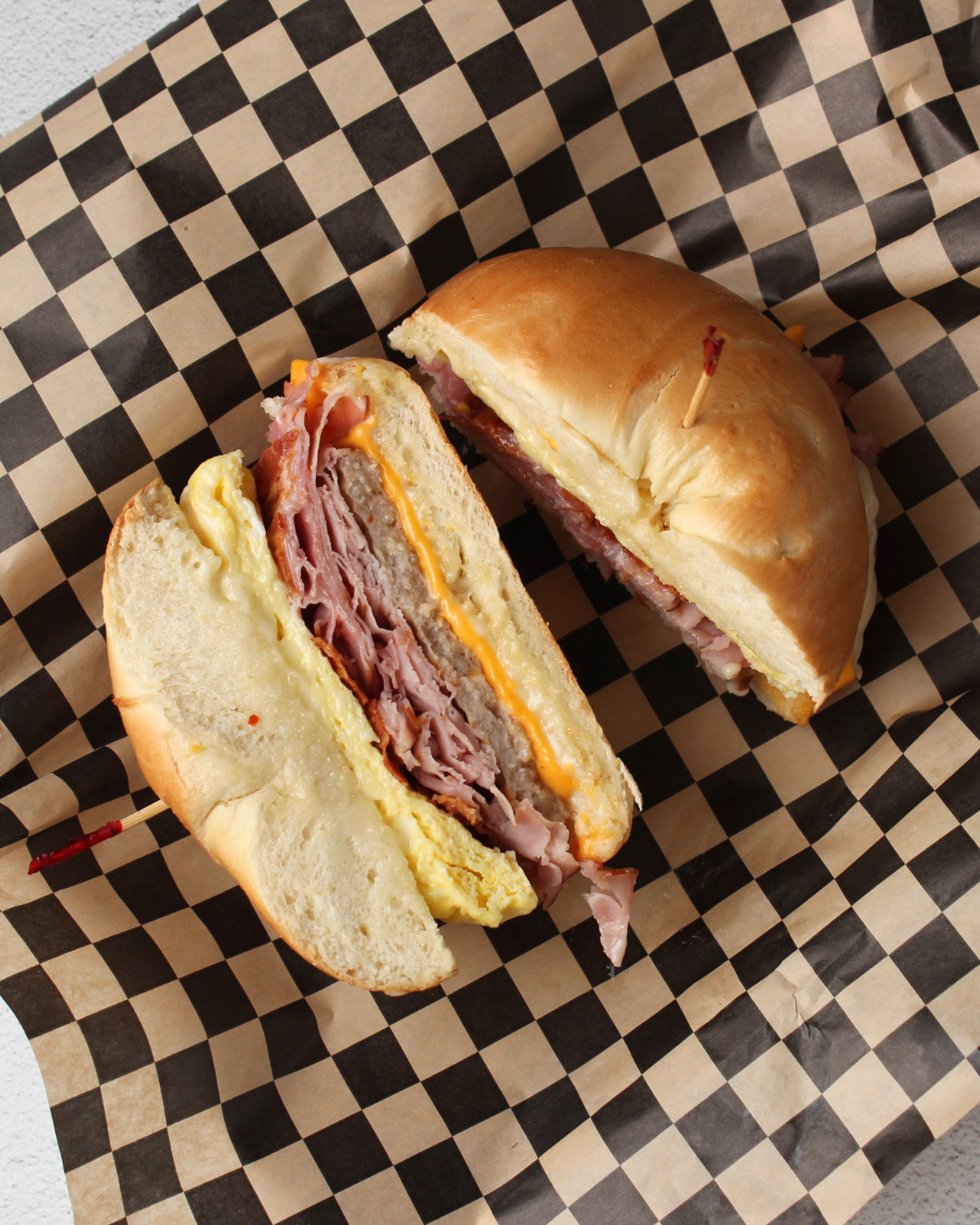 Half of a sandwich on checkered paper, showing layers of ham, cheese, and egg in a bun.