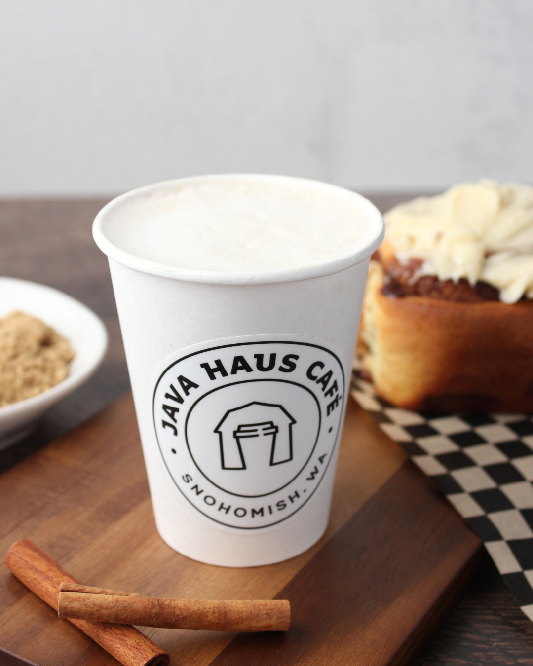 White cup with Java Haus Café logo containing a beverage, cinnamon sticks, and assorted desserts on a wooden tray.