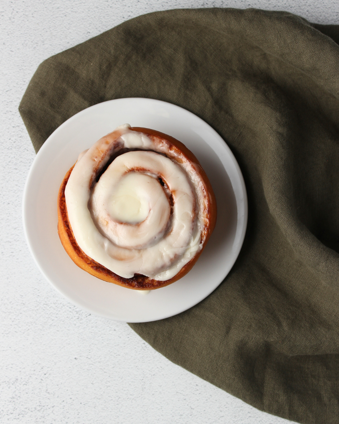 A cinnamon roll with white icing on a white plate, placed on a green cloth.