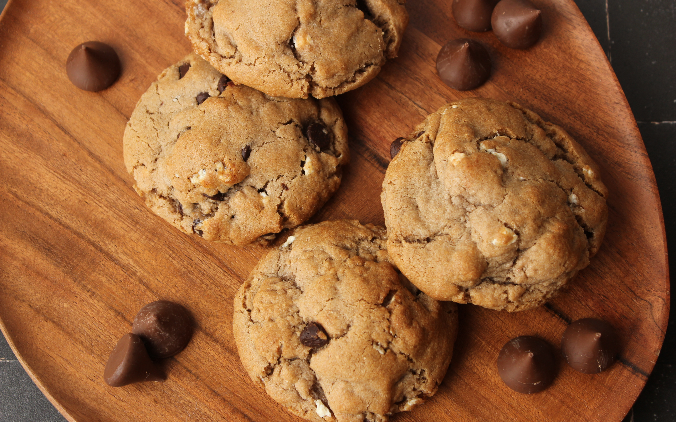 Four chocolate chip cookies with chocolate candies on a round wooden serving board.