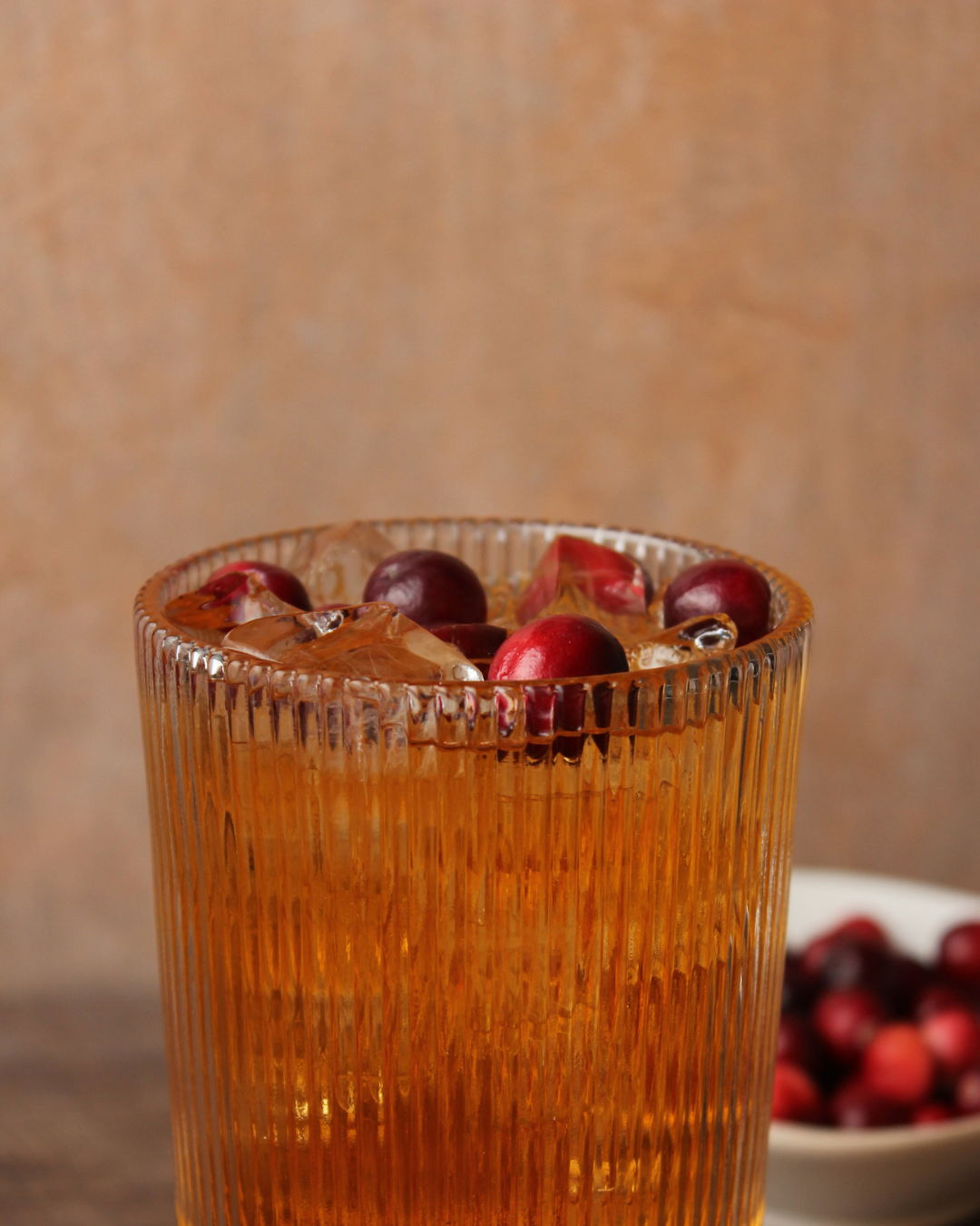 A glass of iced cranberry juice with whole cranberries on top and in a bowl nearby, against a neutral background.